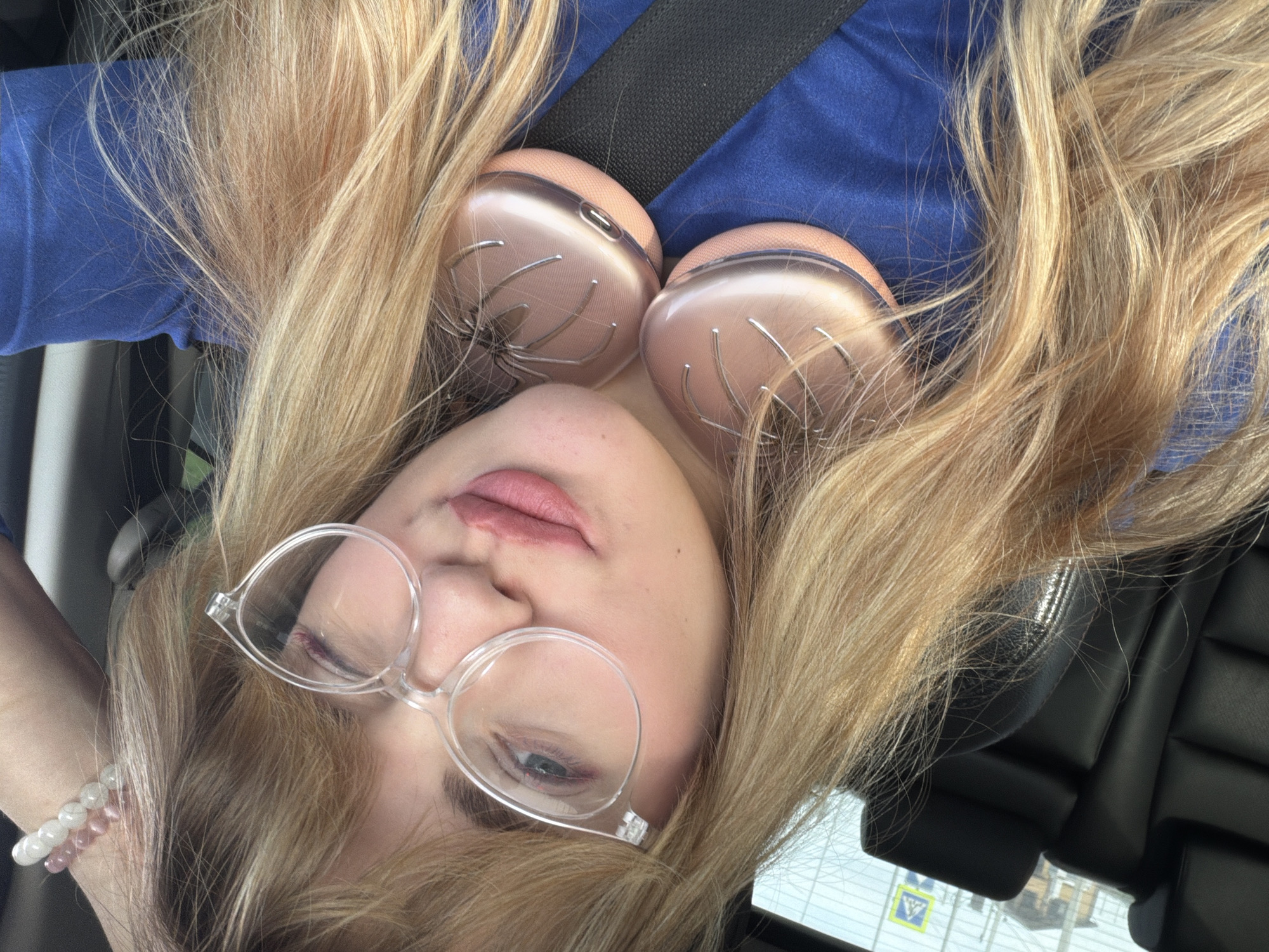 A woman with blonde hair is in a car, wearing a blue shirt and a seatbelt. She has pink headphones on her chest and is wearing clear-framed glasses. She is also wearing a beaded bracelet. A road sign is visible through the car window. Based on the sign, the location is Naberezhnye Chelny, Russia.