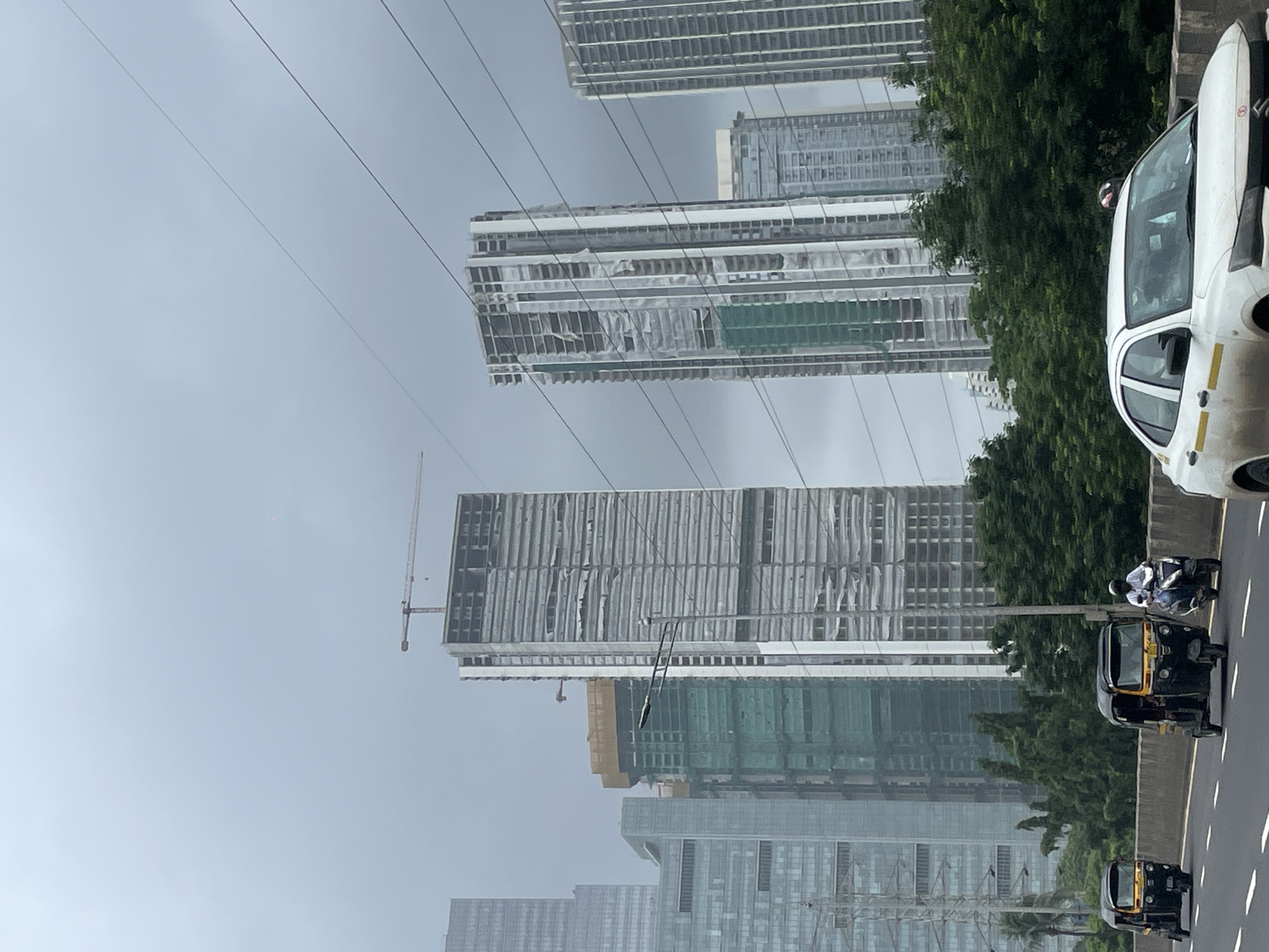 The image depicts a cloudy day in Mumbai, India, likely on a highway. In the foreground, a white car is driving on a road. Behind it, three auto-rickshaws are visible, two black and yellow and one black with a yellow top. A person is riding a scooter.
Dominating the background are several tall, modern buildings under construction. One building, in the center-right, is particularly tall and appears to be a skyscraper in its early stages of completion, with visible scaffolding and construction materials. Another similar building is to its left, also under construction, with green netting covering parts of its exterior. Further left, another completed skyscraper with a patterned facade is visible.
The sky is overcast and gray. Several power lines crisscross the upper portion of the image. Lush green trees line the side of the road, obscuring the bases of some of the buildings. The overall impression is one of urban development and ongoing construction in a dense metropolitan area.