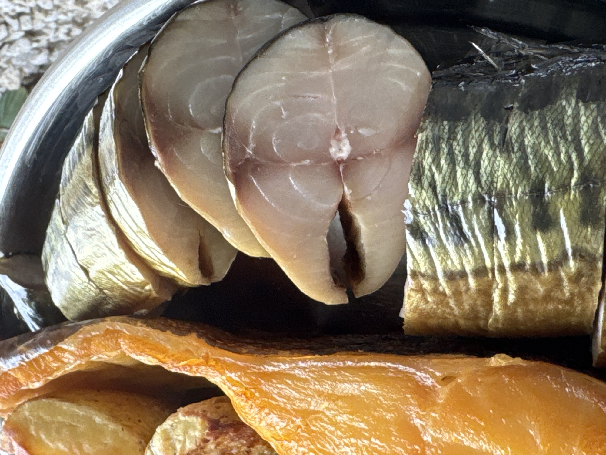 This is a close-up, eye-level shot of smoked fish, specifically mackerel, arranged in a circular silver dish. The fish is cut into thick, circular slices, revealing the pale flesh and darker skin. Some of the fish appears to be whole, with the characteristic wavy pattern of mackerel skin visible. To the left of the fish, a large, golden-orange piece of cured or smoked salmon is visible, its surface glistening. The background is blurred, showing a light-colored, pebbly texture, possibly gravel. The lighting is bright and natural, highlighting the textures and colors of the fish. The image does not depict any people or specific locations within Tallinn, Estonia. It focuses solely on the food presentation.
