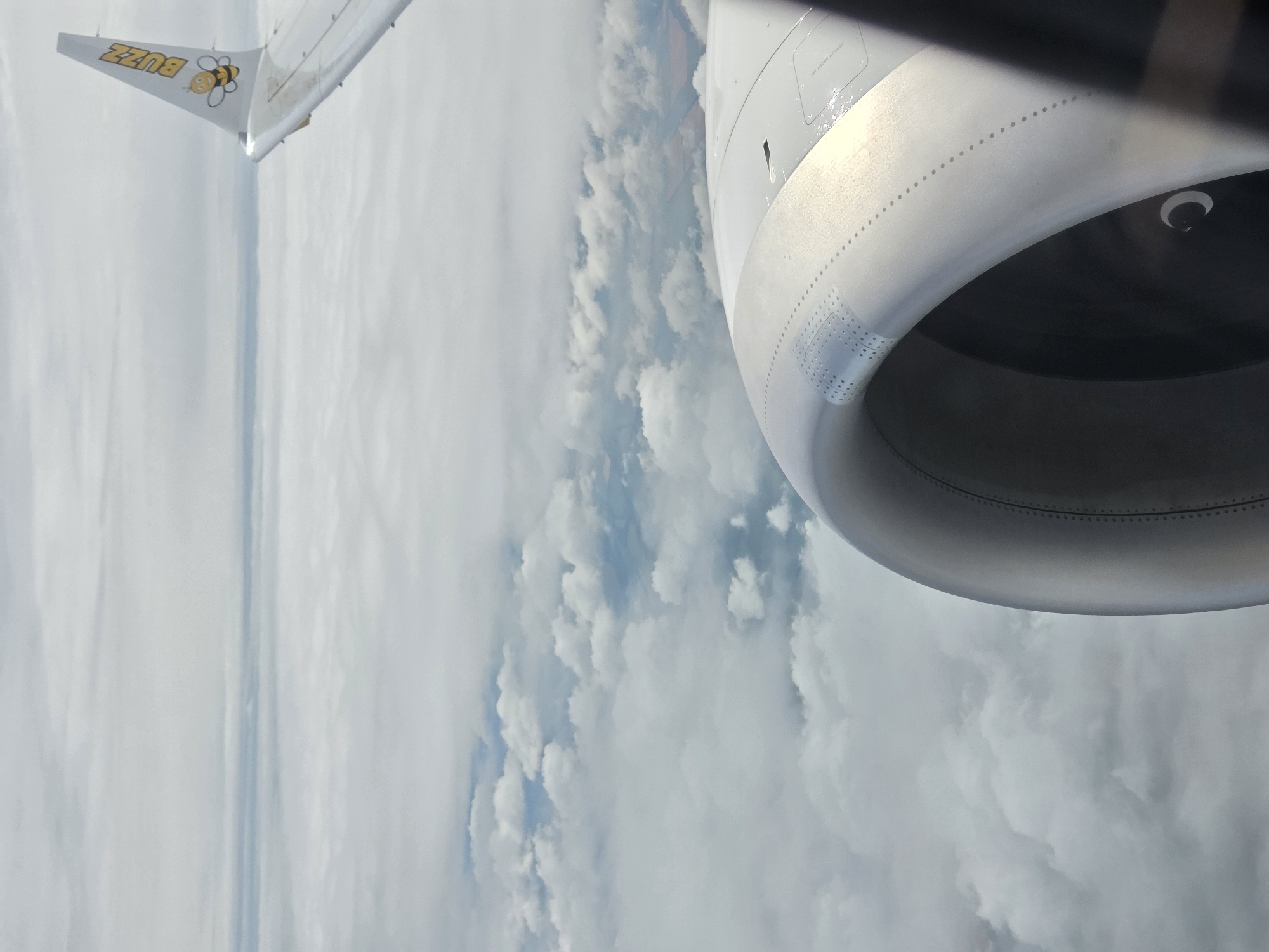 This is an aerial photograph taken from the window of an airplane. The image prominently features the engine nacelle of the aircraft in the foreground, which is metallic and cylindrical in shape. A portion of the airplane's wing is visible on the right side of the frame, featuring the "Buzz" airline logo with a cartoon bee. The background is filled with layers of clouds, creating a soft, hazy atmosphere. The terrain below is obscured by the clouds, but hints of fields and possibly roads can be seen in patches where the cloud cover is thinner. The sky is overcast with a light, diffused light. There are no people or animals visible in the image. The scene depicts a typical view from an airplane during flight, suggesting travel at a high altitude above a cloudy landscape. The location is identified as the city of Orliak, Bulgaria, implying this aerial view is of the airspace above or near that region.