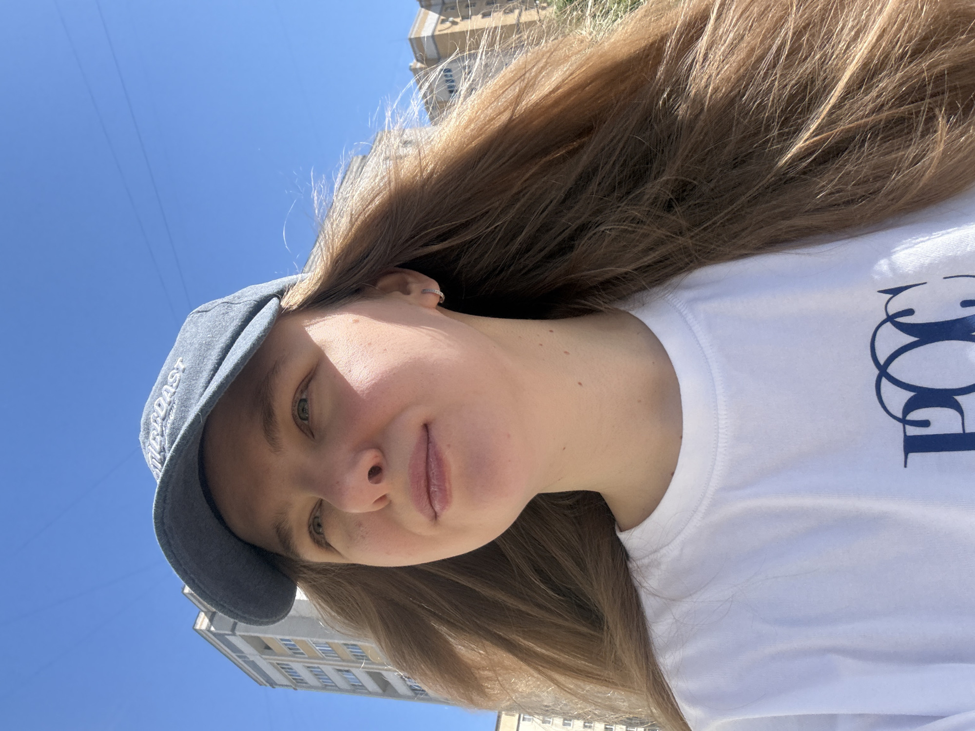 A young woman with long, light brown hair and fair skin is positioned in front of a clear blue sky. She is wearing a light blue denim baseball cap and a white t-shirt with a dark blue graphic on the front. The image appears to be taken from a low angle, with buildings visible in the background. The buildings' architecture suggests a modern urban environment. Based on the information provided, the location is specified as the city of Vladimir, Russia. The woman is looking at the camera with a slight smile.