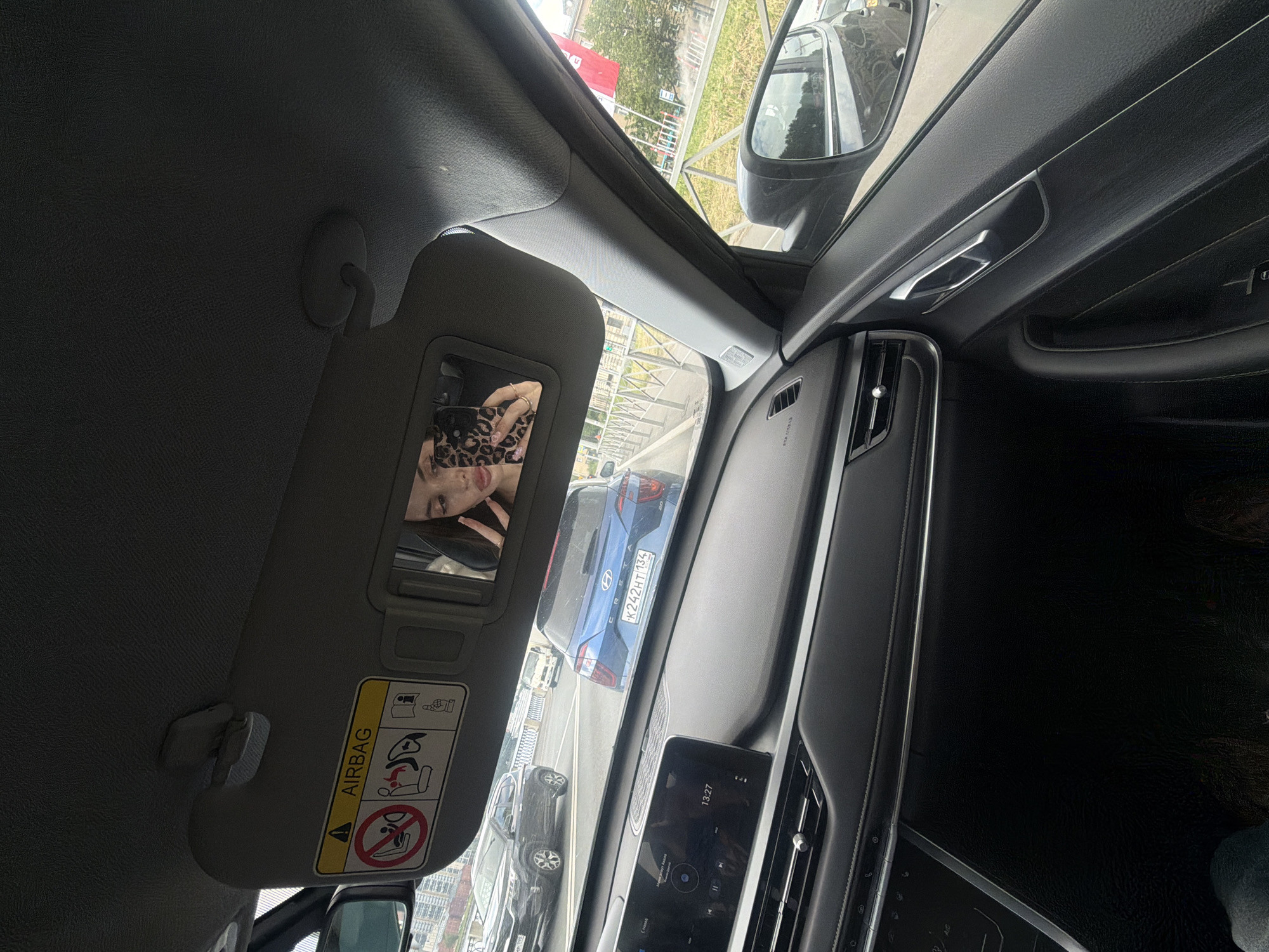 A woman is taking a selfie in the sun visor mirror of a car. She is wearing a leopard print top and has manicured nails. The car is in traffic, with other vehicles visible through the windshield. The dashboard of the car is visible in the foreground, showing a touchscreen display. The exterior environment appears to be a city street with buildings and trees in the background. The sun visor has an airbag warning label.