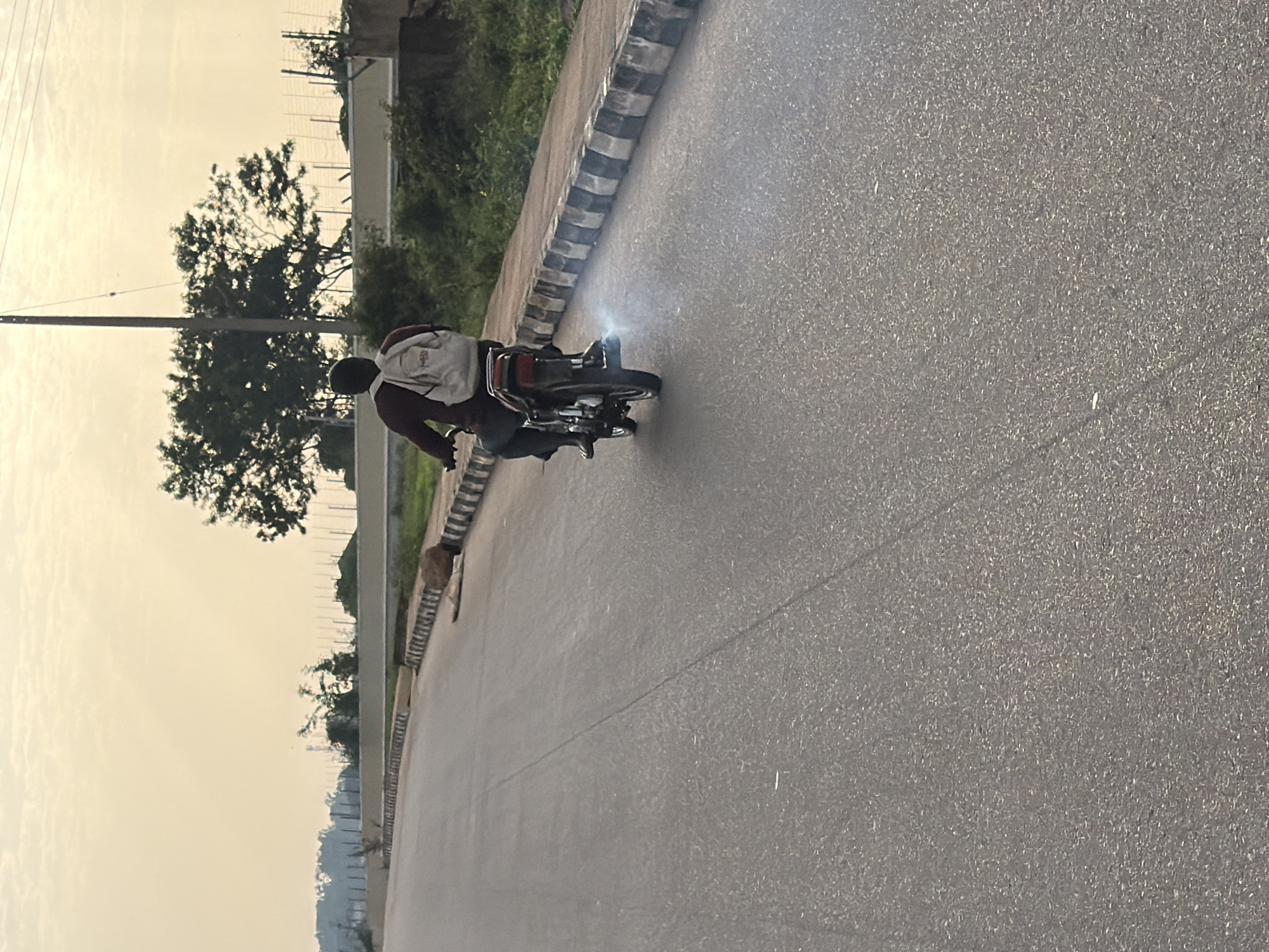 A person wearing a maroon shirt and dark pants, carrying a light-colored backpack, is riding a motorcycle with white smoke emitting from its exhaust. They are on a paved road with a black and white striped curb to their right. In the background, there are trees, a fence, and a utility pole under a hazy sky. There is a large rock on the side of the road. The scene is located on an asphalt road in Gura Top, Nigeria.