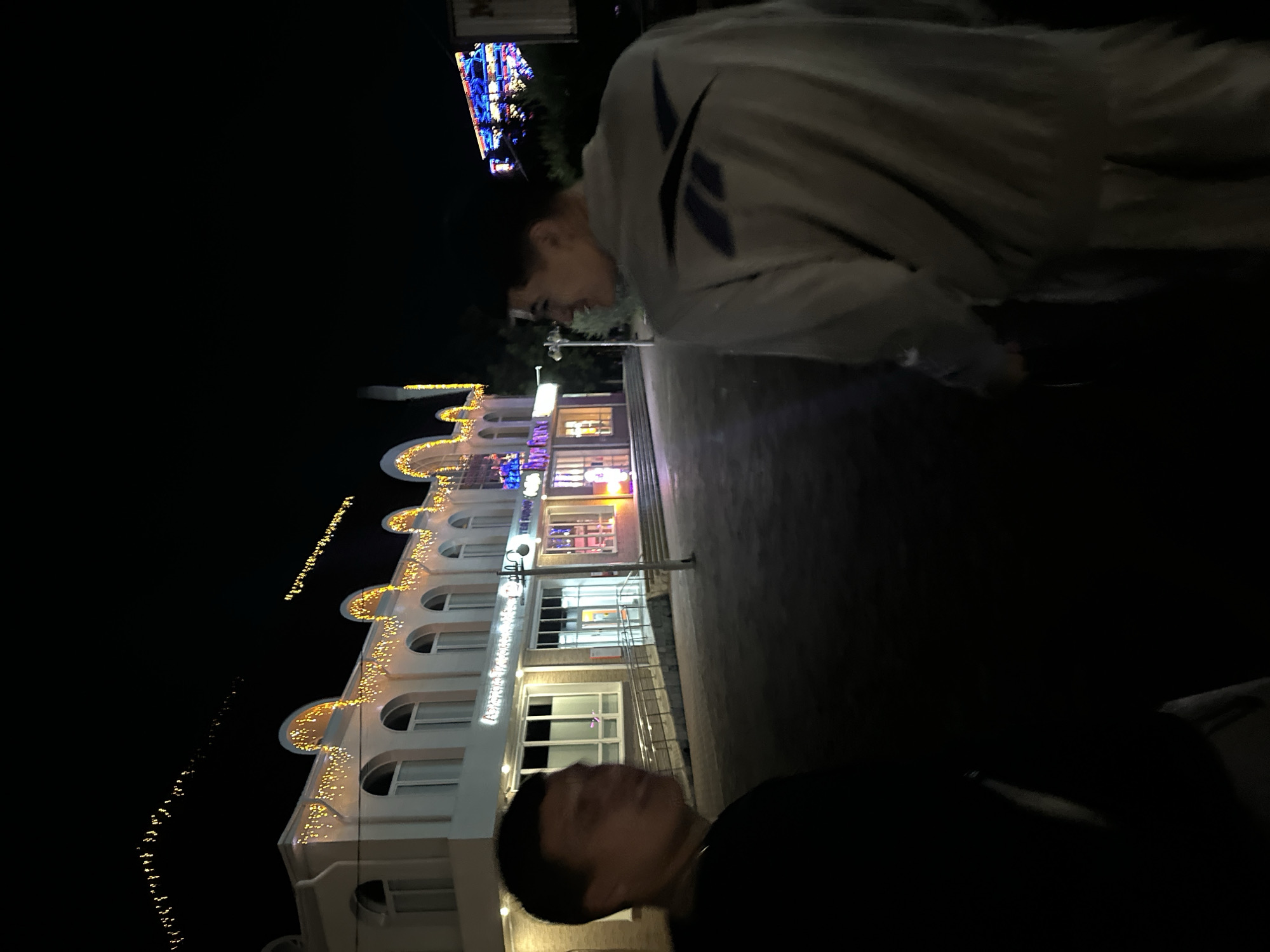 The image shows two people at night, with a building illuminated by festive lights in the background. The person in the foreground is partially visible and appears to be wearing a dark shirt. The second person, further back and to the left, is wearing a light-colored jacket with a dark logo on the sleeve. They are looking down and to the side, with a slight smile on their face.
The building in the background is white with arched windows and is decorated with strings of warm, yellow lights along its facade. Some of the windows are illuminated, revealing interior spaces. There are also colorful lights visible on the upper part of the building, suggesting a festive atmosphere. The sky is dark, indicating it is nighttime. The ground appears to be paved.
In the upper left corner of the image, there is a glimpse of a sign with blue and pink neon lights, suggesting a commercial establishment. There is also some text visible on the building, but it is difficult to read clearly due to the lighting and angle. The overall impression is of a pleasant evening outing in a well-lit urban or commercial area. The activity appears to be a casual gathering or a stroll at night.