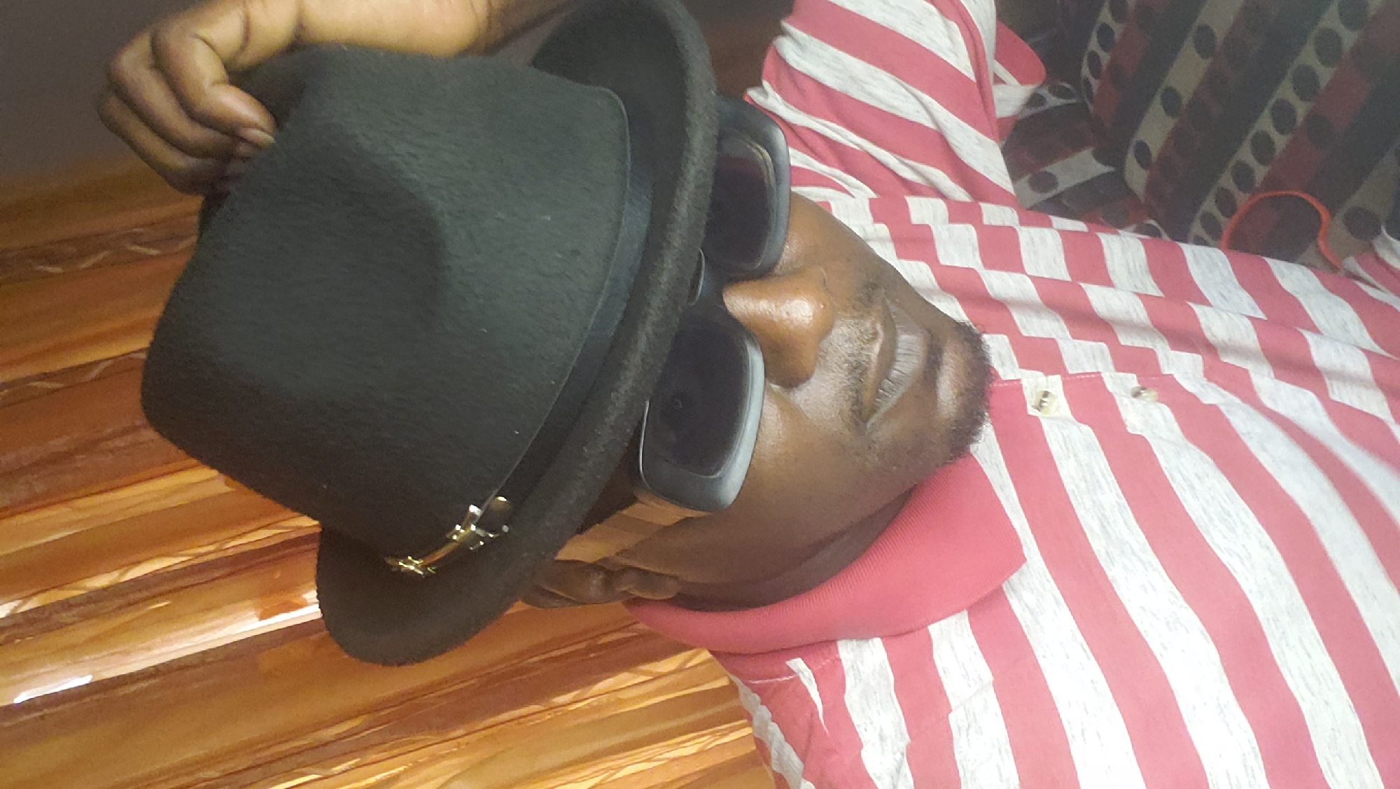 A medium shot depicts an individual lying down, wearing sunglasses and a black fedora hat. The person is dressed in a red and white striped collared shirt, with the collar being a solid red. The background appears to be a bed with patterned linens. The lighting suggests an indoor setting. Information about a specific location like Narayi, Nigeria is not evident in the image.