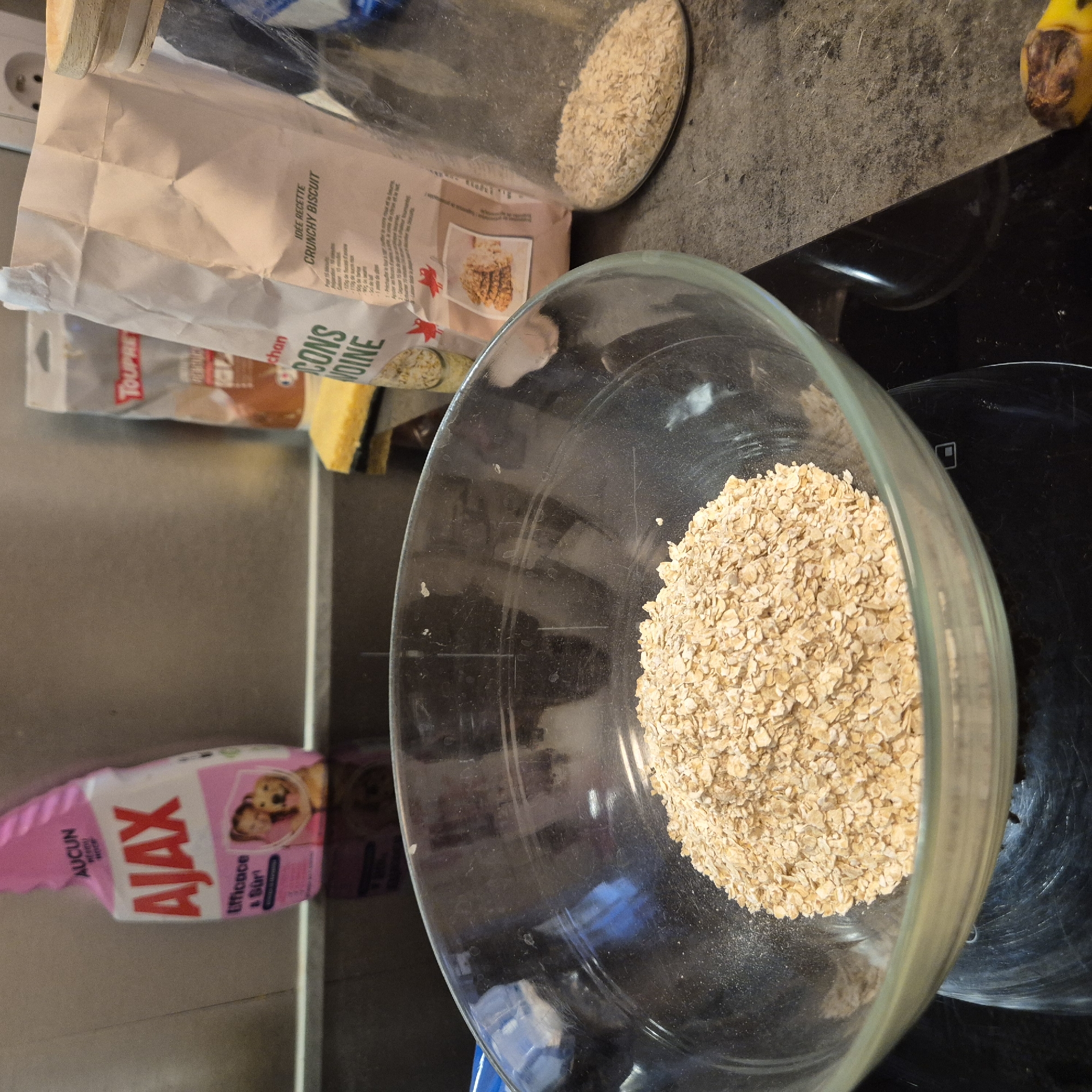 The scene appears to be a kitchen setting, with various items scattered around a kitchen counter. The primary focus is a clear glass bowl filled with rolled oats, sitting on a black surface, possibly a stovetop. To the left, there's a bag of "CRUNCHY BISCUIT" oat flakes, along with what seems to be a bar of soap or cleaning product. A jar filled with oats sits behind the bag, next to the bag of biscuit oat flakes. A bottle of "AJAX" cleaning product with a pink label is situated on a lower surface, possibly a shelf. Based on the label "chan CONS VOINE" on the oat flakes bag, the location is likely Strasbourg, France, as "chan" is part of the Auchan brand, which is a French supermarket chain.