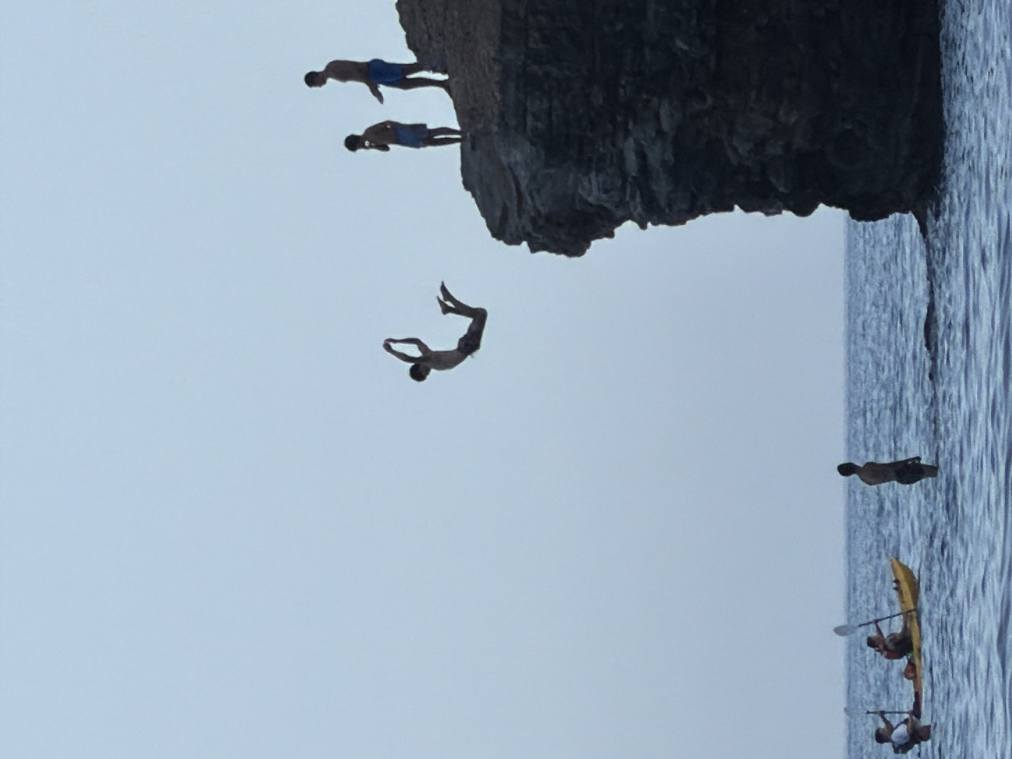 A group of young men are engaged in cliff diving and kayaking on a calm sea with a sheer rock face. In the foreground, two men are in a yellow kayak. One is paddling, and the other is holding a paddle upright. To their right, a single man stands in the water. In the mid-ground, a man is captured mid-air, diving from the top of the rock face. On the edge of the rock, two other men stand, watching the diver. The sky is clear and overcast, suggesting it is either early morning or late afternoon. The scene appears to be taking place at Mogren Beach, a popular spot for cliff diving near Budva, Montenegro.