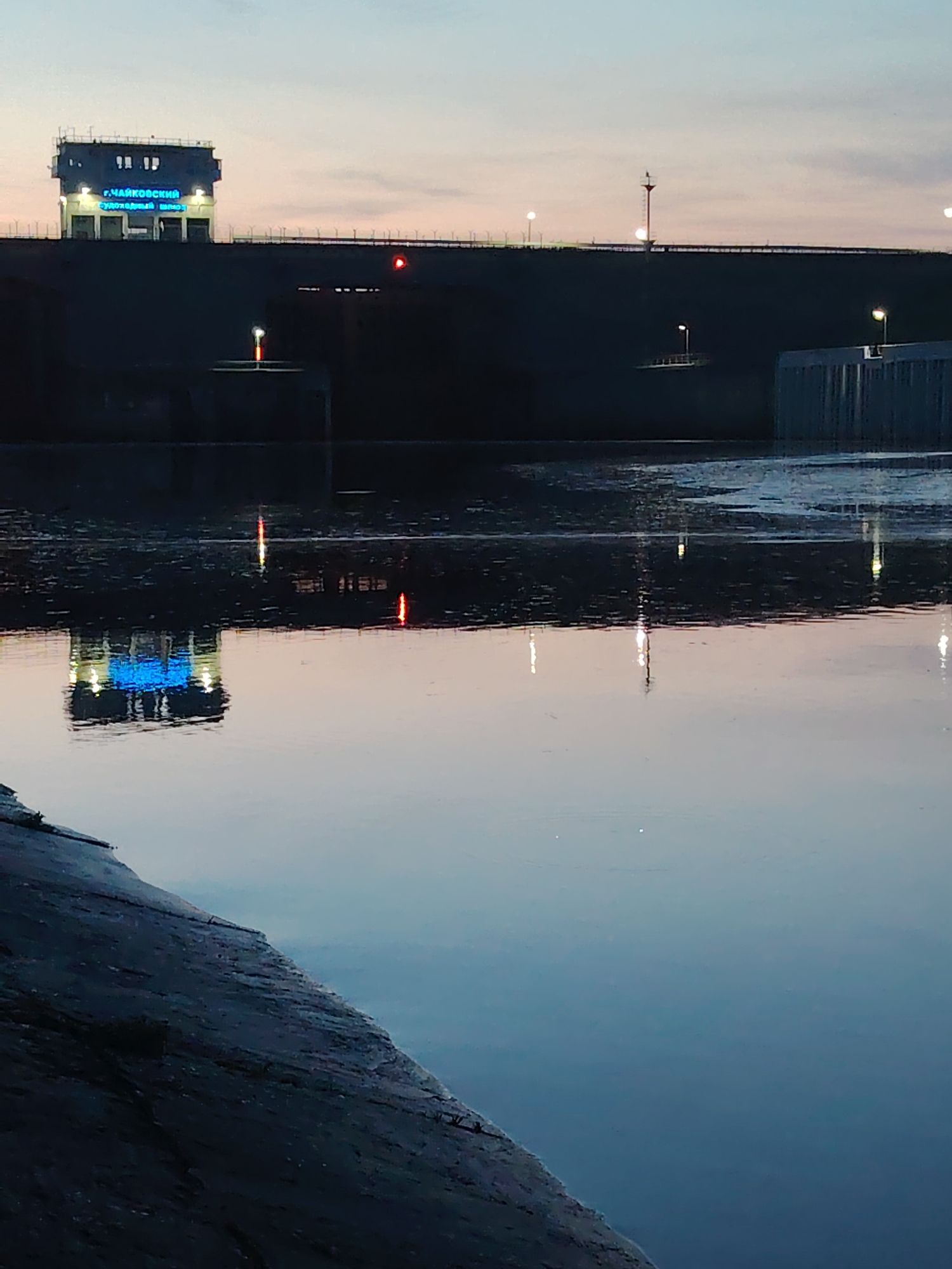 The image depicts the Chaikovsky lock in Chaikovsky, Russia, at dusk or dawn. A multi-level building with illuminated blue signage in Russian sits atop a dark, elevated structure spanning the width of the frame. The structure is also equipped with multiple lights and what seems to be a warning beacon. A body of water reflects the structure and its lights, creating a distorted mirror image. The foreground shows a section of a rocky or paved surface, possibly the bank of the river or canal. The sky is a blend of soft pink and blue hues.