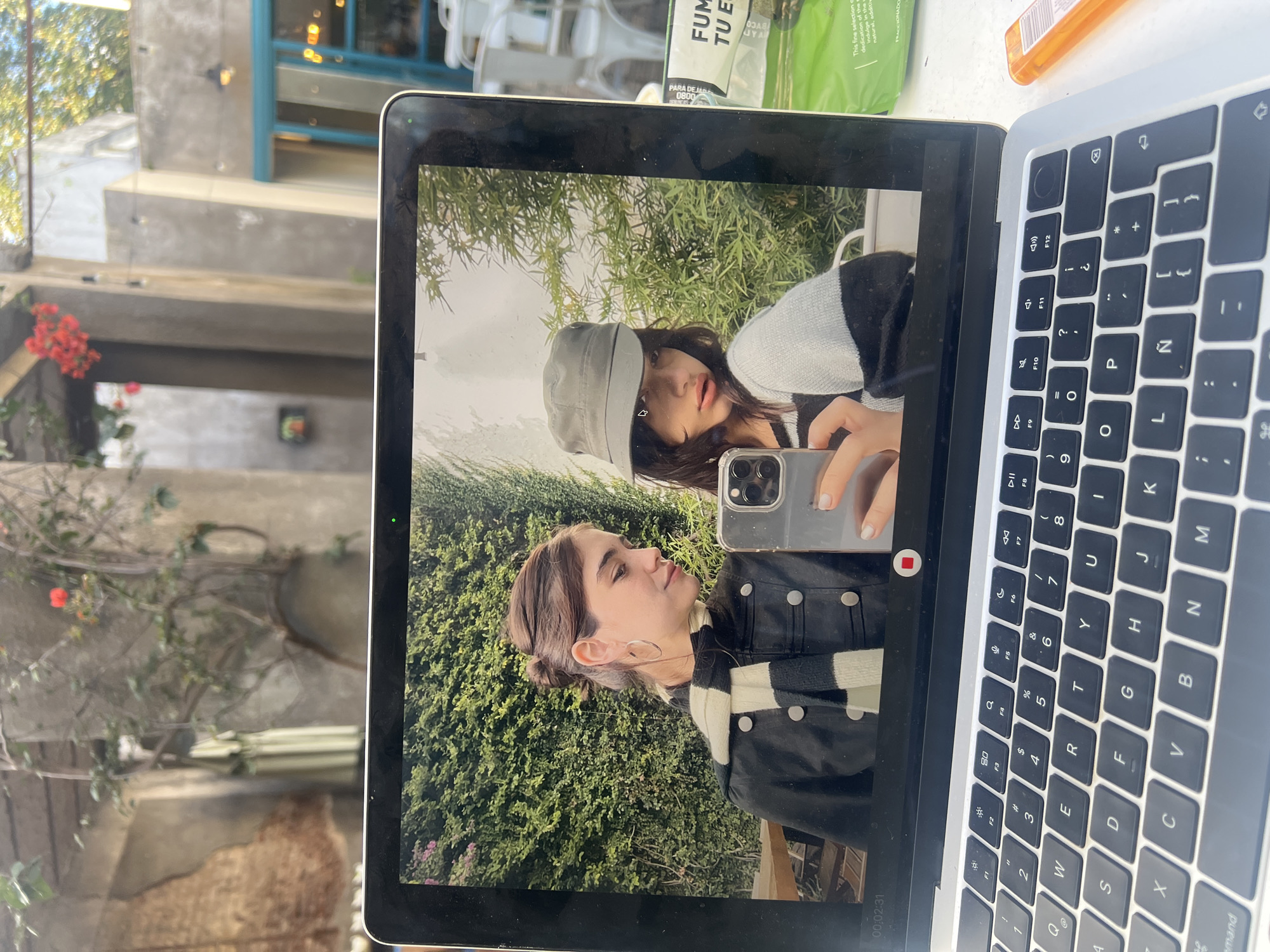 The image captures a MacBook laptop placed on a surface, displaying a video or photo of two young women. The women are shown in an outdoor setting with a dense green leafy background. One woman, wearing a light bucket hat and a striped top, holds an iPhone, seemingly taking a selfie or recording, looking directly at the phone's camera. The other woman, with her hair in a bun and wearing a black jacket over a striped top, is in profile, looking upwards.
The laptop screen displays a timestamp "00:02:31" and a red recording indicator, suggesting a video is being played or a live camera feed. The laptop's keyboard features an "Ñ" key, indicating a Spanish layout, consistent with the Buenos Aires, Argentina context.
In the real-world background behind the laptop, an outdoor environment is visible, including concrete structures, climbing vines with vibrant red flowers (possibly bougainvillea), and a blue railing. To the top right of the laptop, a green and white product package is partially visible with text such as "FUMAR TU E," "PARA DEJA," "0800," and phrases like "This fine selection c," "dedication of th," and "natural, additiv." An orange plastic item, possibly a lighter, is also next to the packaging. The bright, clear lighting suggests it is daytime.