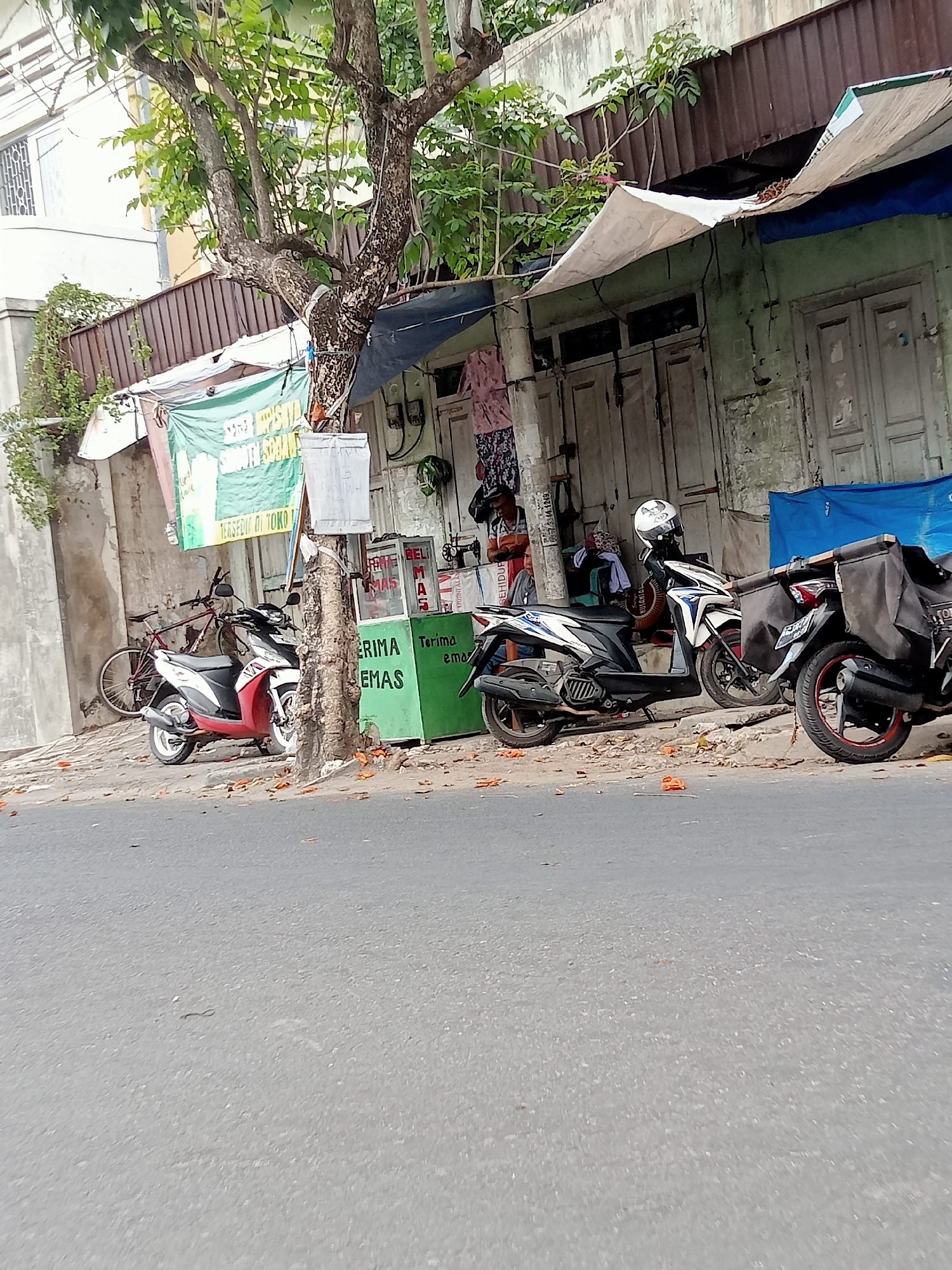 The image depicts a street scene in Tulungagung, Indonesia, characterized by a row of modest storefronts and parked motorcycles. The storefronts appear aged, with weathered wooden doors and faded paint. Tarpaulins are strung up to provide shade or cover from the elements. In front of one shop, a green stand with text is visible, suggesting it might be a vendor's stall. Several motorcycles are parked along the sidewalk, a common sight in Indonesian cities. A bicycle is also leaning against a pole near the storefronts. A tree with sparse foliage grows alongside the sidewalk, its trunk prominent. The street itself is paved, and scattered leaves indicate a potentially dry or windy period. The overall atmosphere suggests a typical urban commercial area in a developing Indonesian city.