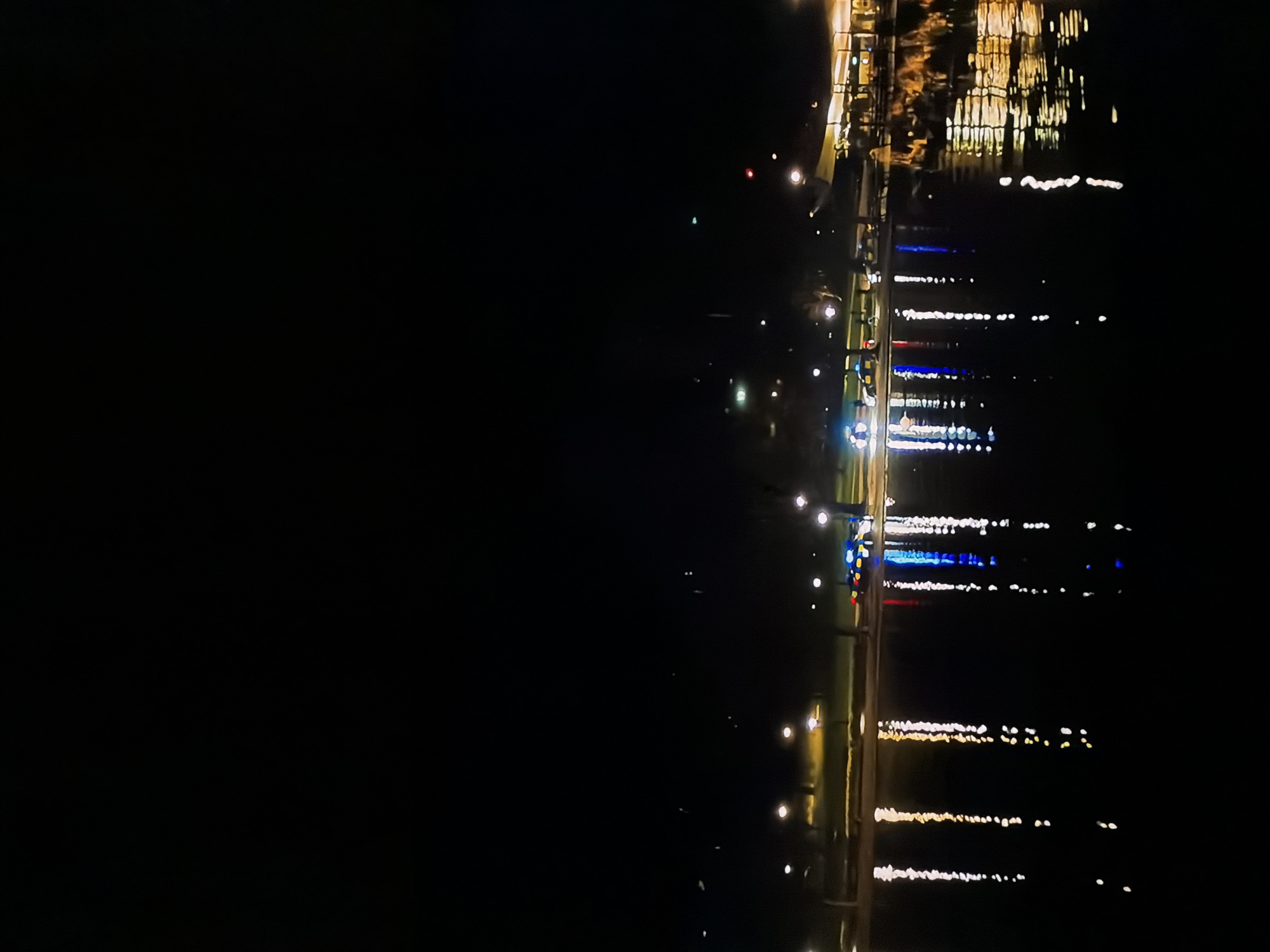 The image, originally oriented vertically but best viewed horizontally, captures a serene night scene of a waterfront or canal in Sofiivska Borshchahivka, Ukraine. The left half of the frame is predominantly dark, indicating open water or an unlit distant shore. The right half reveals a landmass or bank lined with various artificial lights, which cast prominent reflections across the water.
The illuminated bank features numerous light sources, including streetlights, and possibly lights from buildings or decorative installations. These lights create a vibrant display of reflections on the dark water. The reflections appear as elongated, shimmering streaks, primarily in warm yellow and white tones. Distinct clusters of bright blue and some red reflections are also visible, especially towards the middle-right, suggesting a variety of colored lights along the shore. The most concentrated and complex reflections, appearing as a jumble of golden streaks, are present in the upper-right corner, possibly indicating illuminated structures or buildings.
No specific activities or people are discernible in the scene. The water surface appears relatively calm, allowing for clear and defined light reflections. The overall impression is one of peaceful nocturnal ambiance, where the artificial lights of the shore create a striking visual pattern on the water. No text is visible in the image.