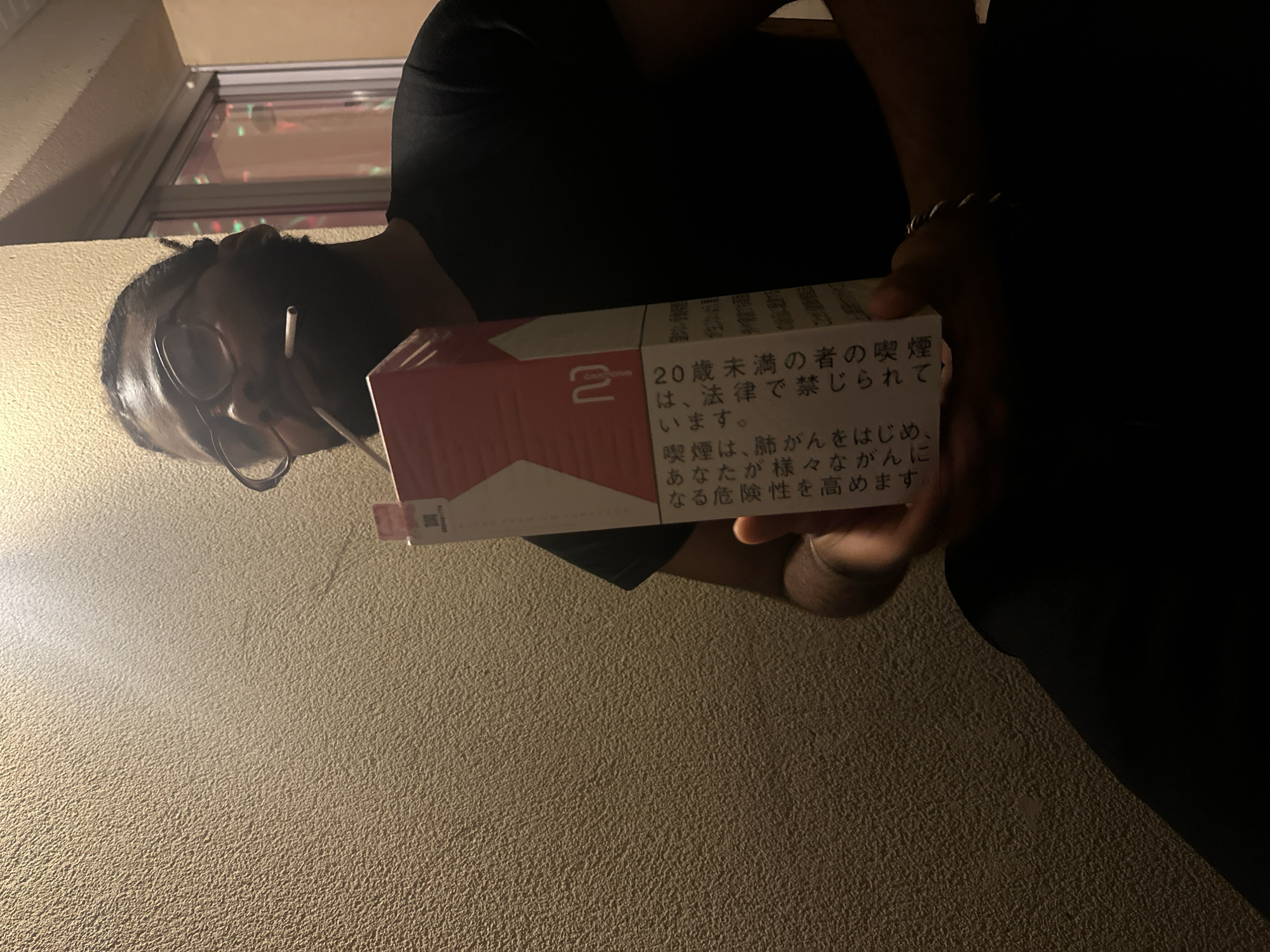The image depicts a person holding a pack of cigarettes, specifically Marlboro 2's, with Japanese text on the side, including a warning about the dangers of smoking for those under 20. The person, who has dark skin, wears glasses and has a cigarette in their mouth. The photo is taken from a low angle, looking up at the person. The environment appears to be indoors with an off-white wall and a window visible in the background. There is no indication that this image was taken in Montreuil, France.