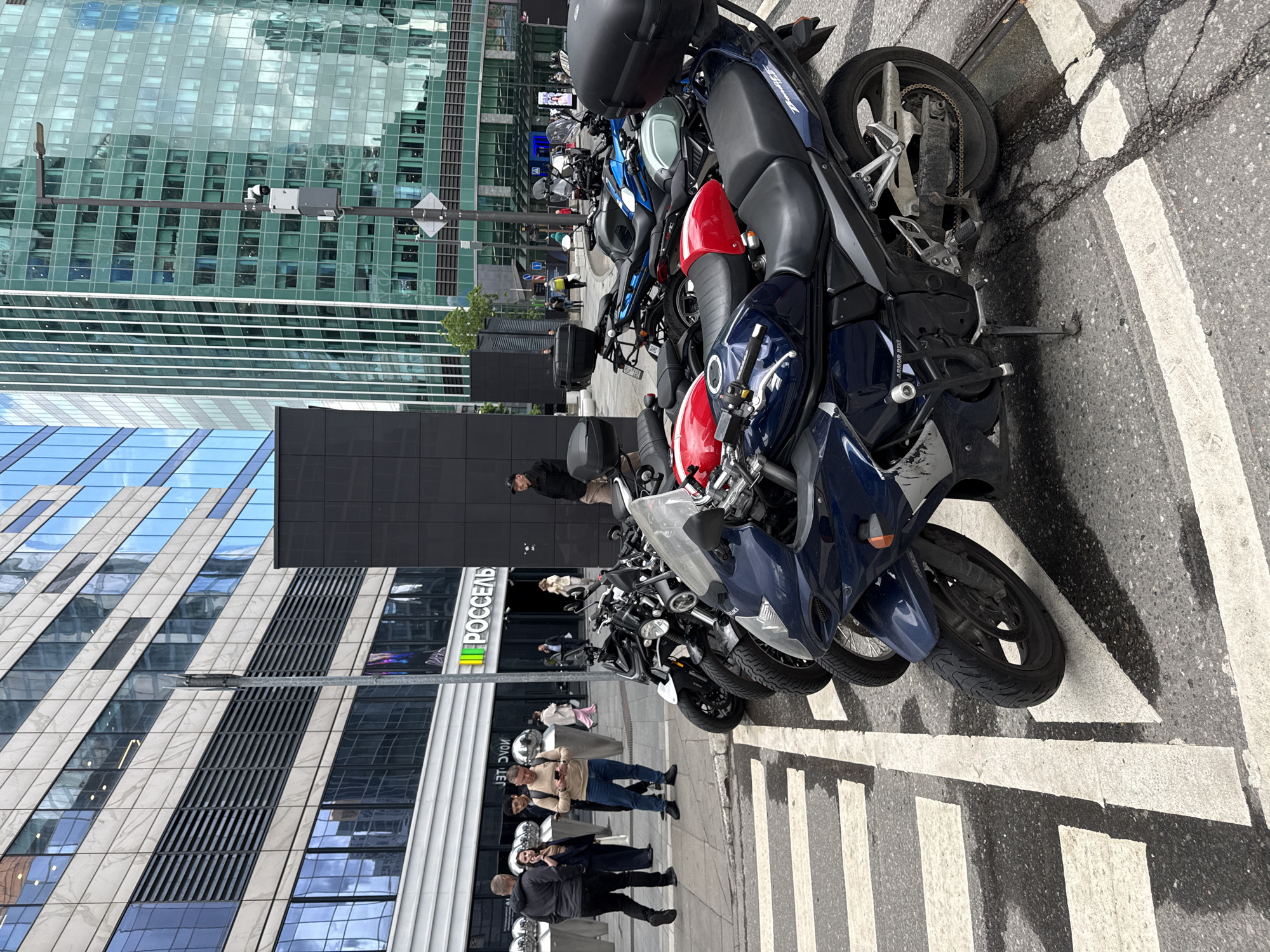 In Moscow, Russia, a street scene unfolds with a row of parked motorcycles in the foreground, predominantly dark blue, red, and black. These motorcycles are positioned on a pedestrian crossing marked with white stripes. Behind the motorcycles, several people are visible on the sidewalk. One man in a beige sweater is looking at his phone, accompanied by other individuals, some of whom are walking away from the viewer. To the left, a modern building with a beige and glass facade stands, featuring a prominent sign with the Russian word "РОССЕАЛЬ". To the right, a tall building with a green glass facade dominates the skyline, with a lamppost equipped with a surveillance camera visible on its pole. The environment appears to be a contemporary urban setting, likely a business district, with clean architecture and well-maintained public spaces.