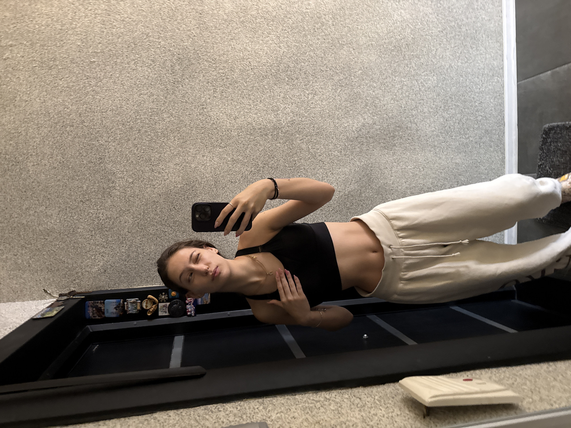 A young woman is taking a mirror selfie in what appears to be an indoor residential setting, possibly in the city of Sofia, Bulgaria. She is wearing a black sports bra and loose-fitting light-colored sweatpants, exposing her midriff. She has a necklace with a cross pendant and a bracelet on her left wrist. Her right hand is holding a smartphone, capturing the reflection. In the background, a dark door is visible, adorned with several small souvenir magnets. The wall behind her has a textured, speckled finish. A light-colored object, possibly an intercom or a wall-mounted light fixture, is mounted on the wall to her left. The flooring appears to be a combination of dark tiles and a dark rug.
