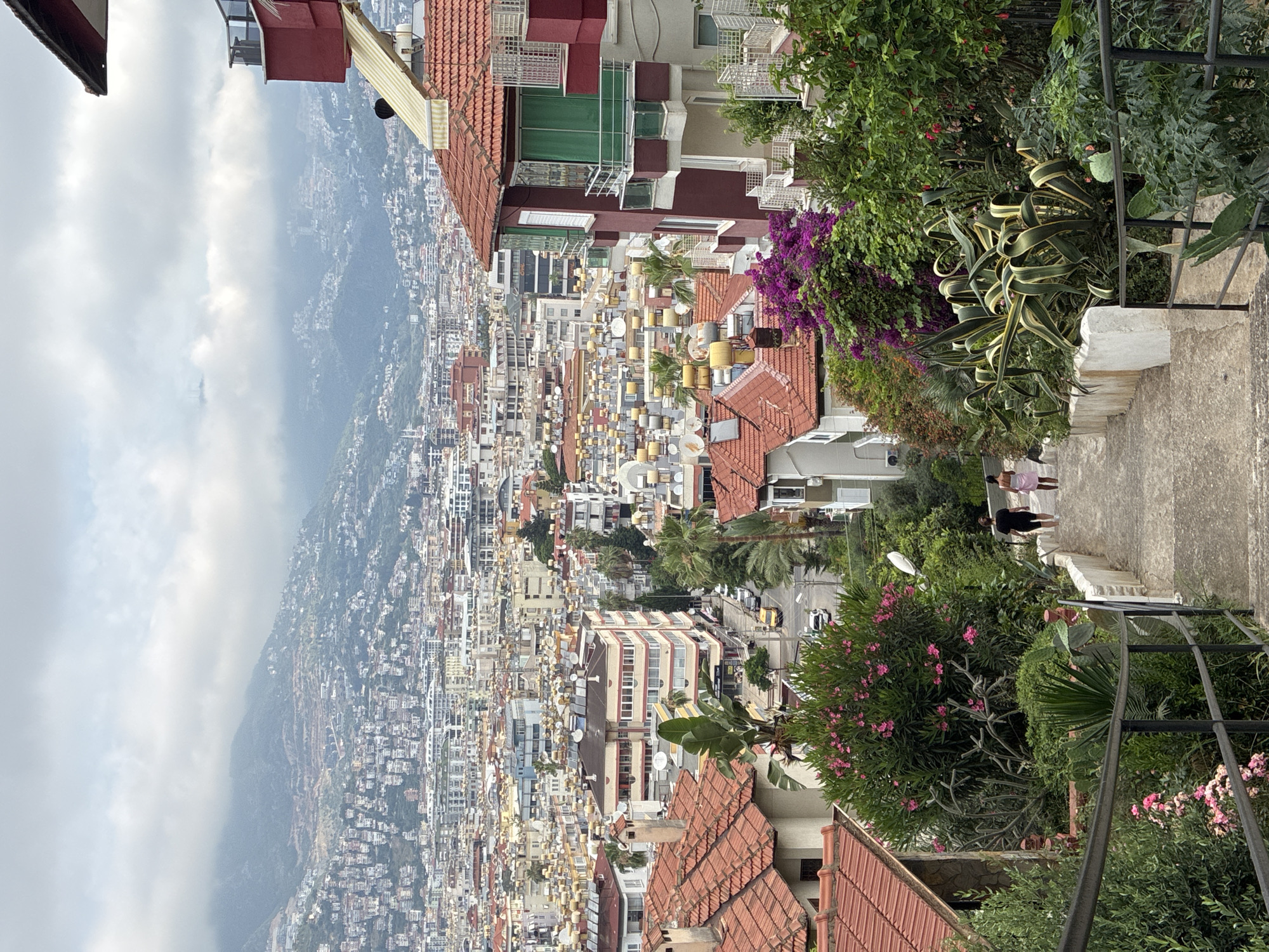The image depicts a view of Alanya, Turkey, looking down a concrete staircase towards a dense urban landscape. In the foreground, lush green vegetation with pink and purple flowers lines the staircase, which is bordered by a metal railing. Two individuals, one wearing a black t-shirt and shorts, and the other a pink dress or sarong, are walking down the stairs.
Further down, a street is visible with a few parked cars. The city unfolds beyond, characterized by a multitude of buildings with red-tiled roofs and solar water heaters prominently displayed on many of them. In the background, rolling hills are covered with more densely packed buildings, extending towards mist-shrouded mountains under an overcast sky. The architectural style of the buildings suggests a Mediterranean or coastal urban environment.
