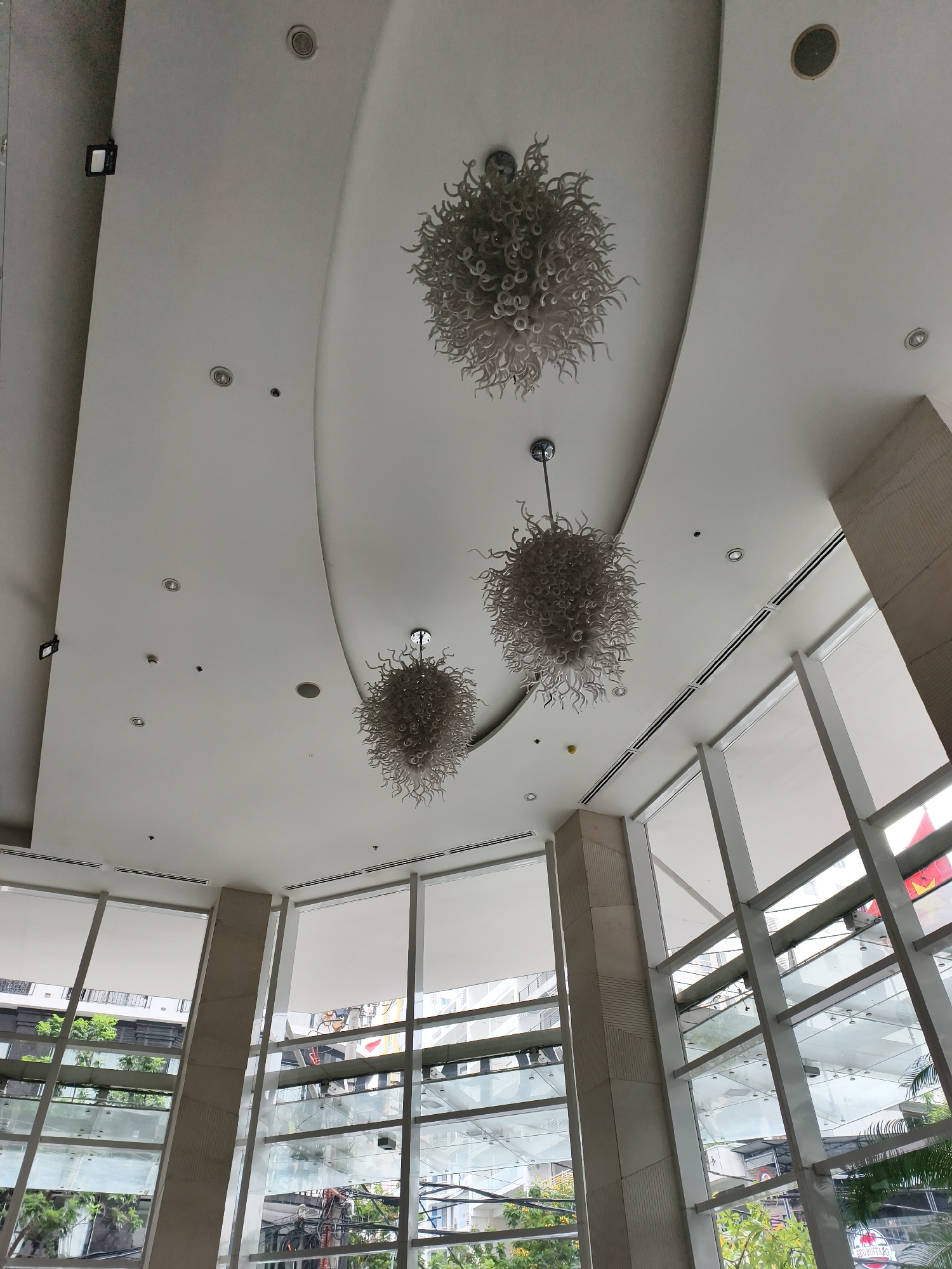 The image captures the interior of a modern building, likely a lobby or atrium, characterized by its high ceilings and extensive use of glass. The perspective is looking upwards and outwards, showcasing architectural details.
The ceiling is predominantly white, featuring a prominent, elegant curved element that spans across the frame. Embedded within the ceiling are numerous recessed spotlights, circular speaker grilles, and small sensors or smoke detectors. Hanging from the ceiling are three distinctive, spherical chandeliers. These light fixtures are composed of a multitude of small, curled, or wavy white/light grey elements, giving them a unique, organic, and textured appearance, somewhat resembling a cloud or coral formation. Air conditioning vents with dark grilles run along the edges where the ceiling meets the walls.
The building's exterior walls are made up of large, multi-paneled windows with light-colored frames, allowing natural light to flood the space. White roller blinds or shades are partially drawn at the top of some window sections. Through the glass, an urban environment is visible, including other buildings, lush green trees, and overhead power lines.
Several thick, light-colored pillars or columns, some with subtle geometric textured patterns, provide structural support and delineate the window sections.
No people or specific activities are discernible in the image. The scene suggests a bright, clear daytime, given the natural light pouring in. A faint red sign is visible through one of the windows, with the text "RED BUFFALO" partially legible. The overall impression is one of spaciousness, modernity, and sophisticated design.
