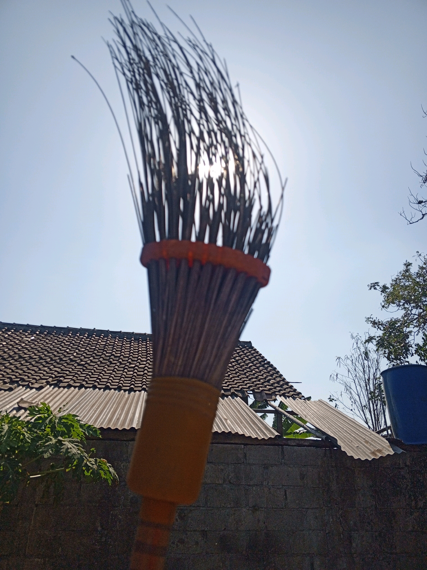 In the foreground, a broom is held up, its bristles silhouetted against a bright sky. The sun is partially visible through the broom's sparse strands. The broom handle is a dull orange color. In the background, a tiled roof of a building is visible, with corrugated metal roofing underneath. A dark brick wall forms the base of the structure. To the right, a large blue water tank and the branches of a tree are seen. The scene appears to be outdoors, under a clear, sunny sky, likely in a residential area within the city of Bantul, Indonesia.