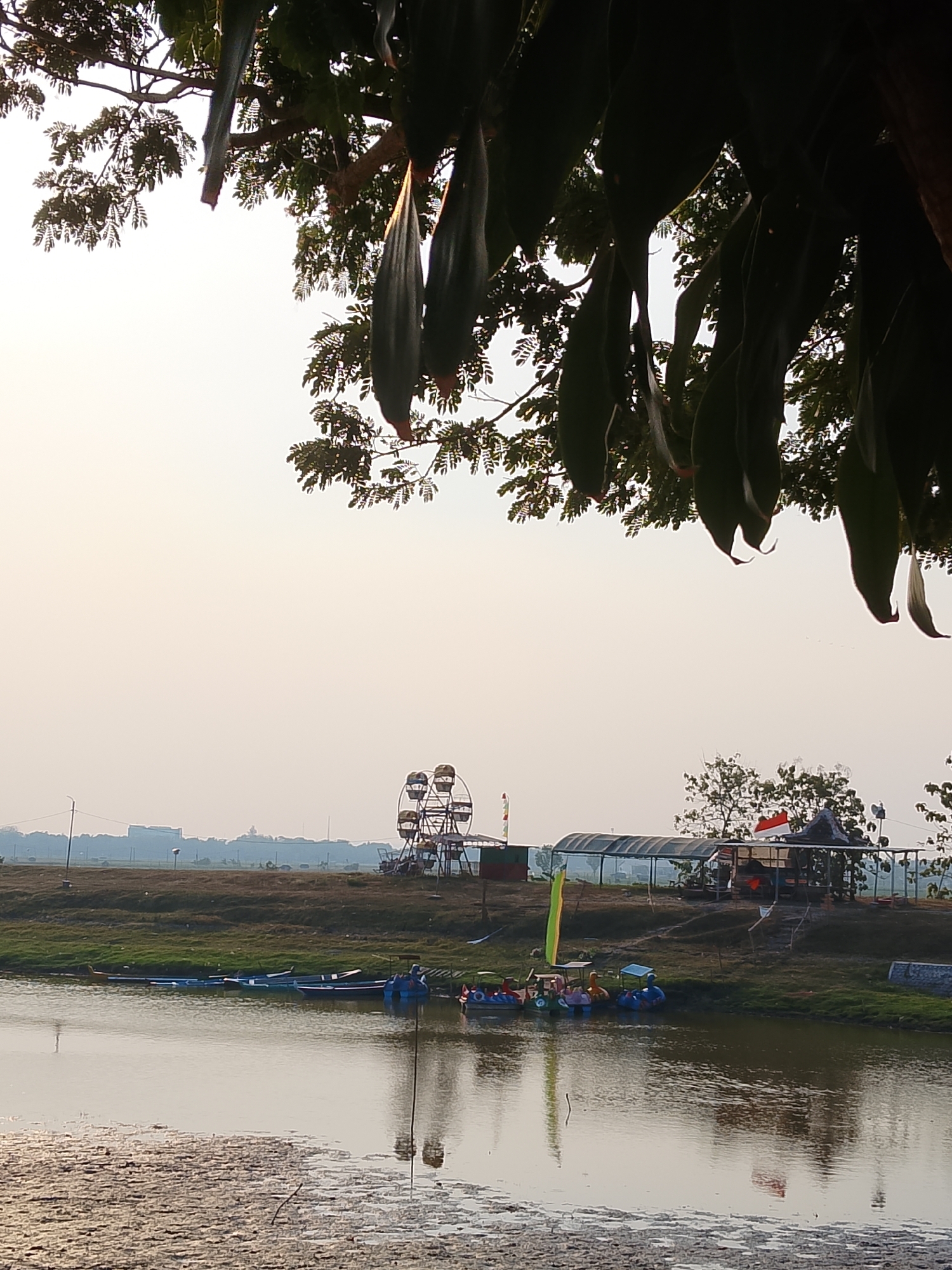 This is a medium-shot, eye-level, outdoor photograph taken in the city of Ngadrin, Indonesia. The foreground is dominated by a body of water, reflecting the sky and trees. The water's edge is lined with mud and vegetation.
In the middle ground, a grassy bank separates the water from a flat, open area. Several small boats are moored along the bank, including what appear to be pedal boats. Further back, a Ferris wheel is visible, suggesting an amusement area. A covered pavilion with a dark roof and open sides stands near the Ferris wheel, adorned with a red and white Indonesian flag. Several trees are scattered across this area.
In the background, a few buildings and structures are faintly visible on the horizon. The sky is overcast and a pale, washed-out white, indicative of either early morning or late afternoon light. The branches and large, dark leaves of a tree hang down from the top of the frame, partially obscuring the sky. There are no people clearly visible in the scene.