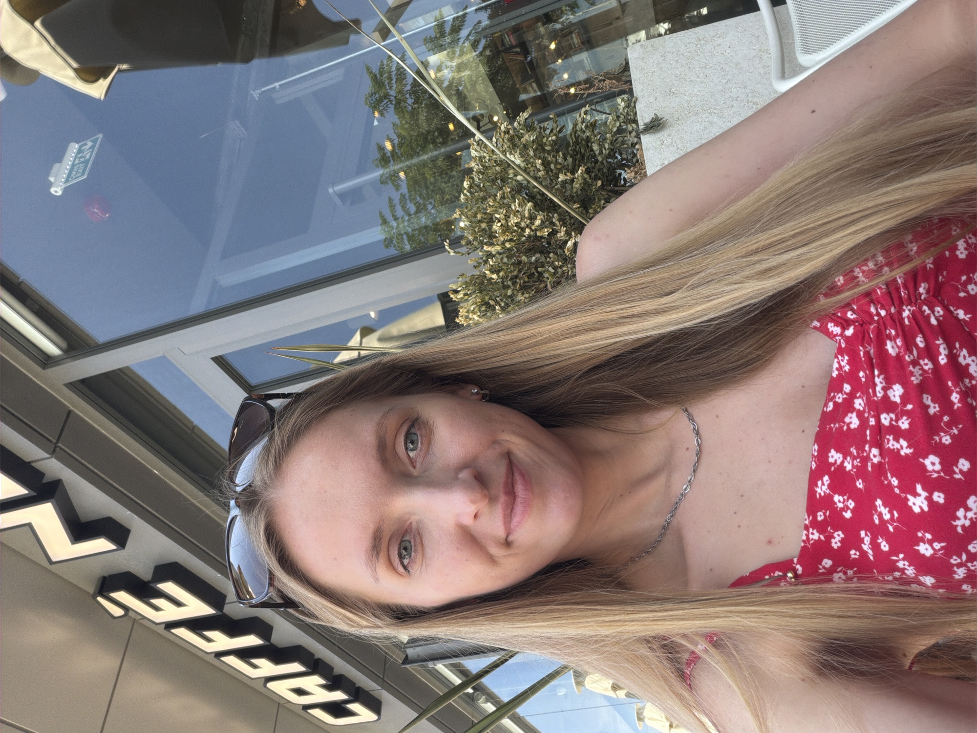 A young Caucasian woman with blonde hair and blue eyes is taking a selfie outdoors. She is wearing a red dress with a white floral pattern and a silver necklace. Sunglasses are perched on her head. Behind her, a sign that reads "CAFFE" is visible, and the reflection of trees and buildings can be seen in the glass facade of the establishment. The lighting suggests it is daytime. The background suggests an urban cafe setting, possibly in Kuşadası, Turkey.