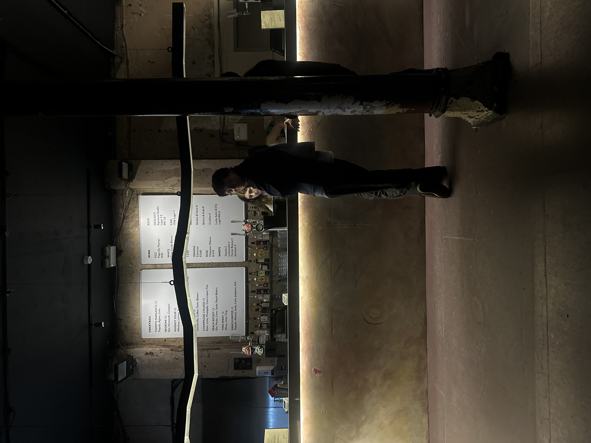 Here is a detailed description of the image:
The image shows the interior of a bar or pub with a dimly lit, industrial-chic aesthetic. Dominating the foreground is a thick, dark metal support structure, possibly a pillar or beam, that runs horizontally across the frame. Patches of corroded or worn paint are visible on its surface.
Behind this structure, the bar area is visible. A menu board displaying drinks lists is on the left. The bar is staffed by two individuals who seem to be a male and a female, with the male standing closer to the foreground. The lighting in the bar is warm, with a strip of vertical light along the back wall providing a focal point. Various bottles are stocked behind the bar.
The general atmosphere appears relaxed and inviting.
Given the drinks offered as listed on the menu, I am unable to tell the location in the city of London, United Kingdom.
