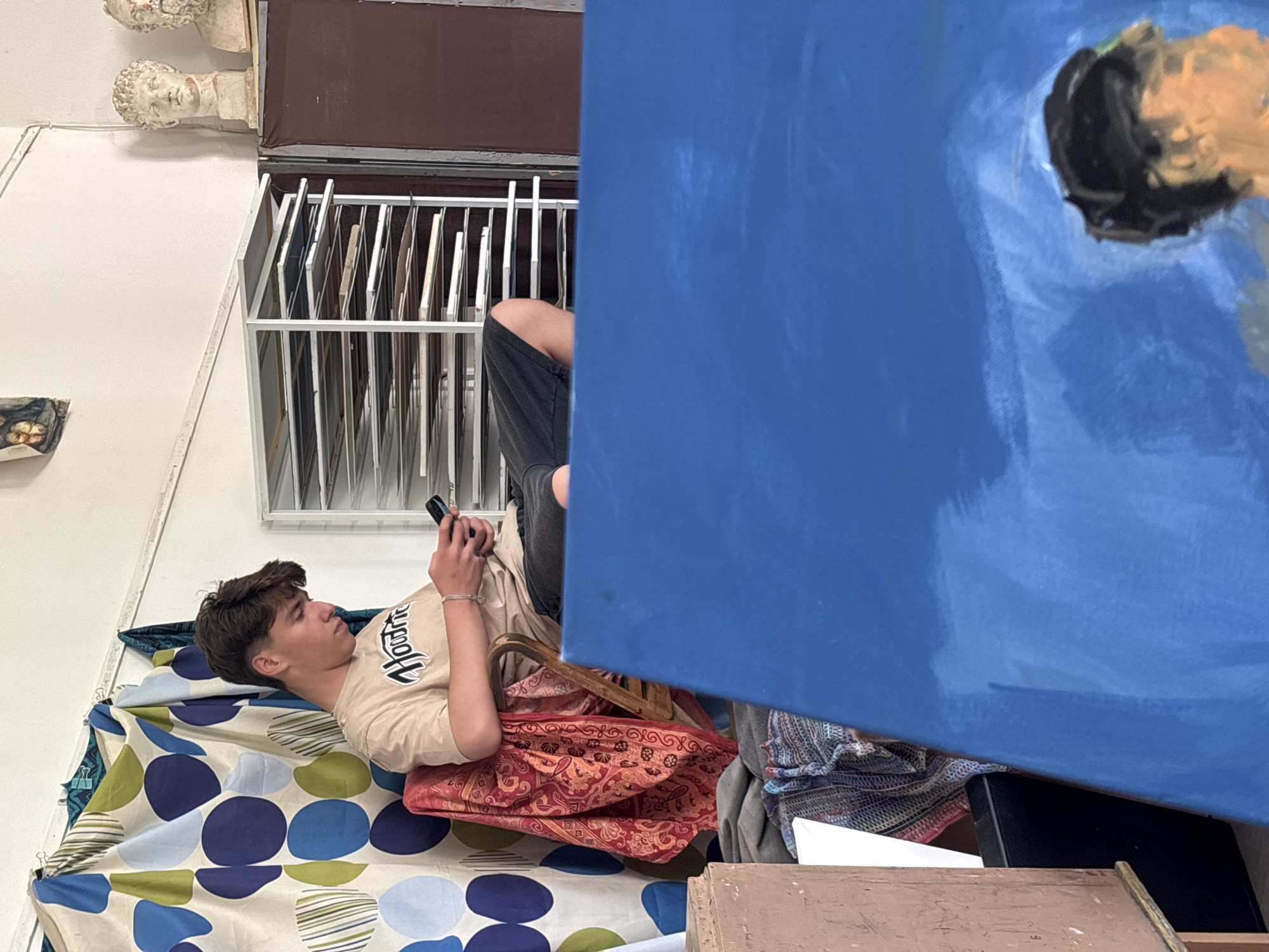 The image depicts a young man lying on a patterned surface, possibly a blanket or cloth, with a red cushion beneath his head and upper body. He is dressed in a beige t-shirt and dark pants, and he is holding a cell phone in his hands, seemingly looking at it. Behind him, there is a metal rack filled with various canvases or frames. To the left, on the wall, there are plaster busts mounted. A large, blue-painted canvas partially obscures the view. It is not possible to confirm whether this image was taken in Troyan, Bulgaria.