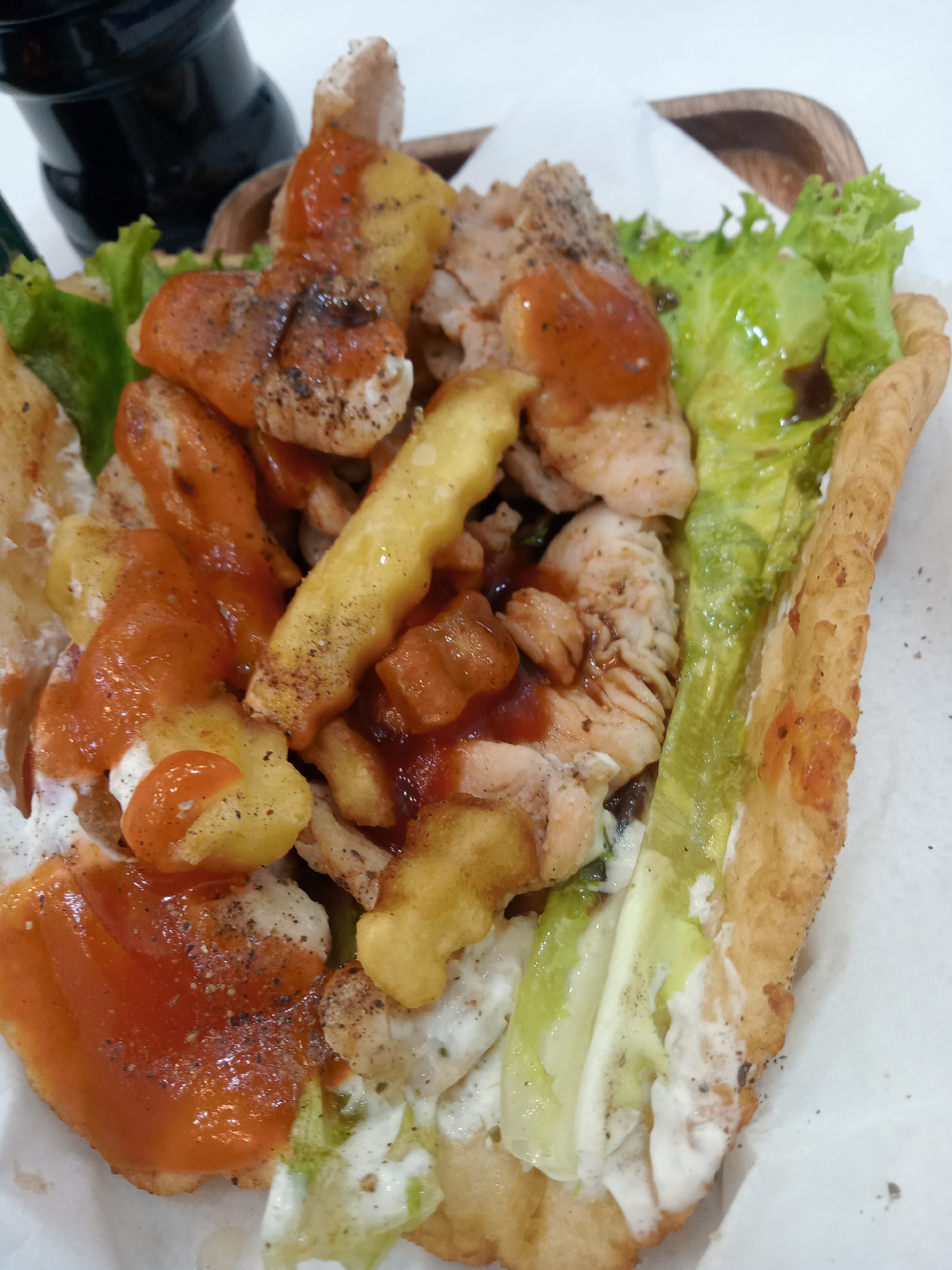A close-up shot depicts a generously filled pita or flatbread sandwich, served in a wooden basket lined with white paper. The filling consists of sliced chicken or pork, french fries, lettuce, and various sauces. Red and white sauces are prominently visible, along with a dark sauce drizzled on the lettuce. Pepper is sprinkled on top. The sandwich appears to be located in a restaurant in Nha Trang, Vietnam, suggested by the style of food. A black pepper grinder is partly visible in the upper left corner of the frame. The setting is indoors, likely on a white table, given the uniform white surface beneath the basket.