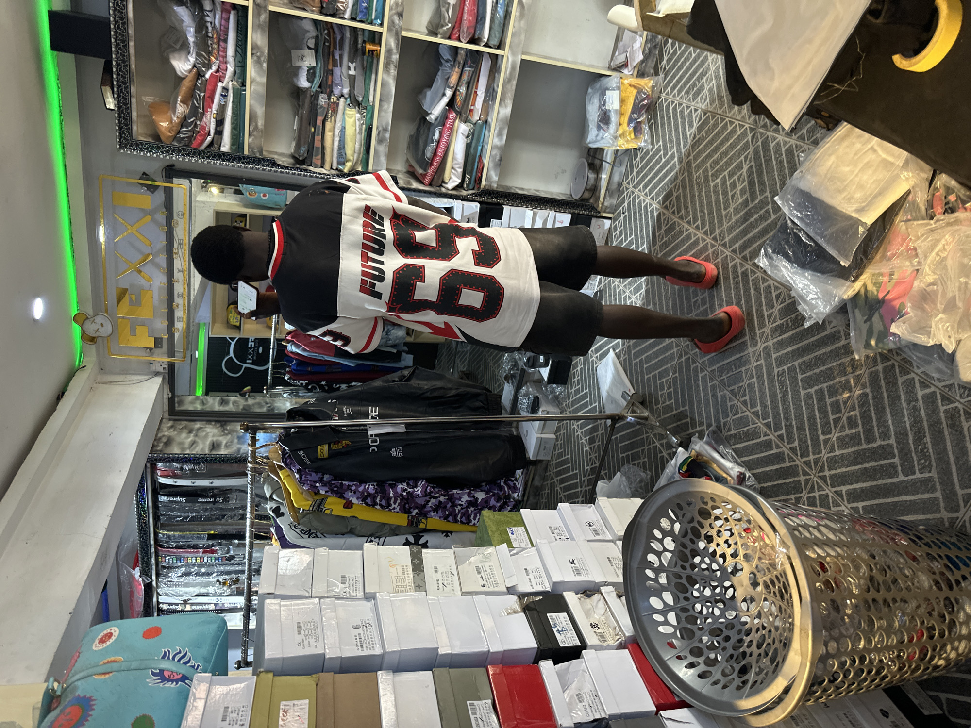 A full shot shows the interior of a clothing store in Bukuru, Nigeria, with a young Black man standing in the center facing away from the camera, holding a phone. He wears a white and black long-sleeved shirt with red trim and "FUTURE 85" printed on the back, and dark shorts. Red slides are on his feet. To his left, stacks of white boxes are piled high. To his right, a tall shelving unit is filled with folded t-shirts and other clothing items. Behind him, clothing racks hold various garments, including jackets and shirts. A large, silver laundry basket filled with items sits in the foreground to the left. The store's floor is tiled with a dark gray and black geometric pattern. A sign above the man's head displays "FEXXI COLLECTION" in a golden font against a clear background, illuminated by green LED lights. Additional green lights are visible along the top of the wall. The environment appears to be a retail space with a focus on streetwear.