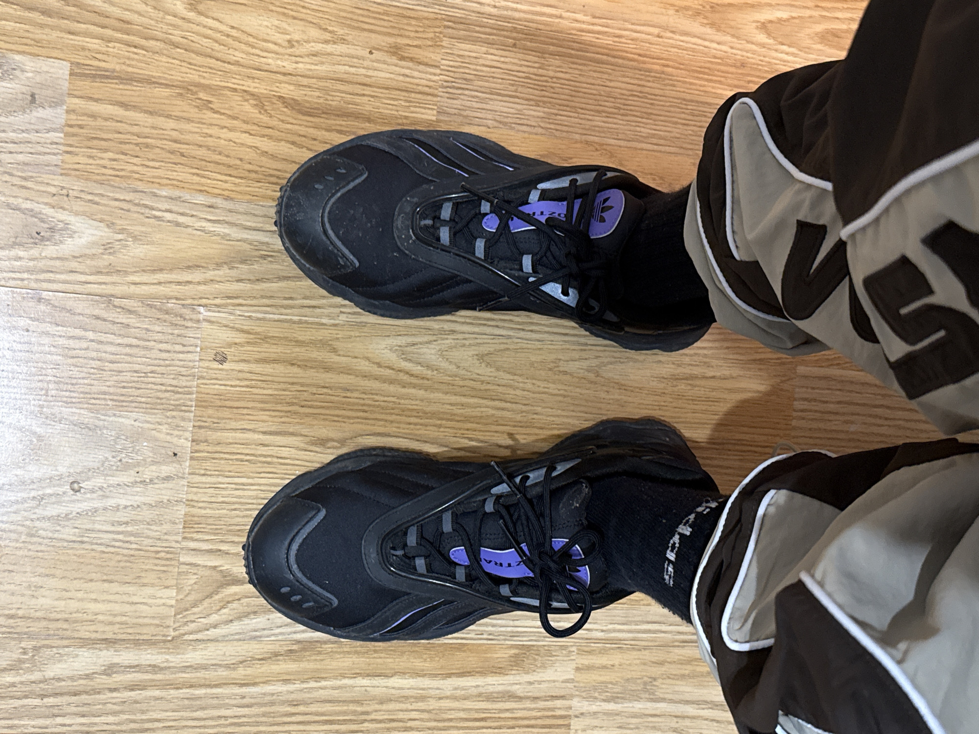 The image, captured from a top-down perspective, showcases a person's lower legs and feet, clad in athletic wear, standing on a light brown wooden floor. The setting appears to be indoors, potentially a home or gym, with the given location context of Kardzhali, Bulgaria.
The subject is wearing black sneakers, characterized by a chunky sole and a black upper with distinctive purple accents on the tongue and reflective grey details around the lacing system. The tongue of each sneaker features an Adidas Trefoil logo in purple and partially visible text "OZTRAL" (likely part of a model name such as "OZWEEGO" or "OZTRAIL"). The shoes exhibit minor signs of dust or wear.
Beneath the sneakers, black socks are visible, displaying the "adidas" brand logo and text on both. The person is also wearing track pants or joggers that are dark brown or black, featuring prominent light grey or beige panels and white piping along the seams. Large, dark, partially obscured lettering, possibly "VNS" or similar, is visible on the grey panel of the right leg.
The overall scene presents a casual, sporty ensemble. The activity depicted is simply standing, allowing for a clear view of the footwear and lower attire. The lighting suggests an indoor environment.