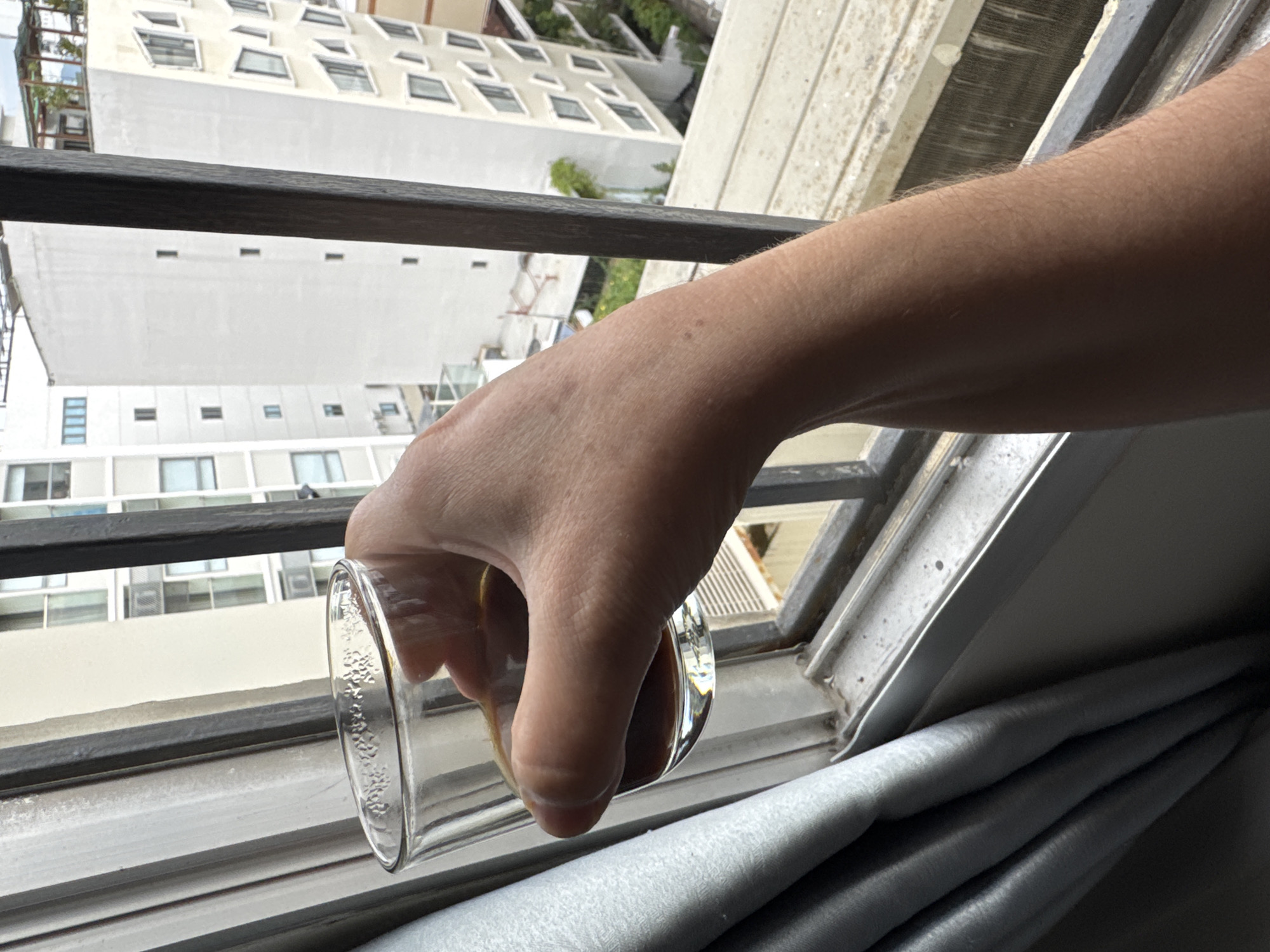 A person's hand is holding a clear glass of dark liquid near a window overlooking a city. Buildings of varying heights and architectural styles are visible outside the window. The window has metal bars. The scene suggests an indoor perspective looking out at an urban environment. The presence of a glass of liquid implies a moment of relaxation or consumption. The backdrop of buildings is characteristic of a city in Vietnam.