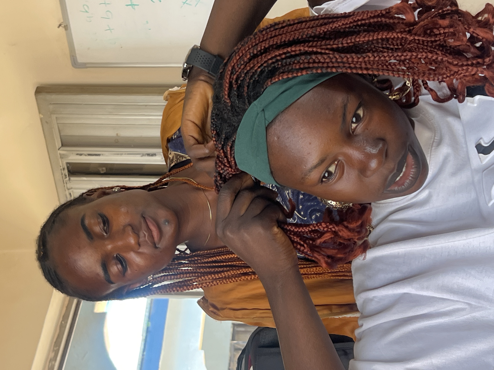 In Jos, Nigeria, two young Black women are engaged in braiding hair. The woman in the foreground, wearing a white t-shirt and a green headband, is seated and looking forward, her mouth slightly open as if speaking. Her hair is braided in thick, copper-colored strands. The woman in the background, standing, is in the process of braiding the hair of the woman in the foreground. She is wearing an orange garment and her expression is focused on her task. The environment appears to be indoors, possibly a classroom or a home, with a whiteboard visible in the background with some mathematical equations written on it. A window with a view of the outdoors is also present.