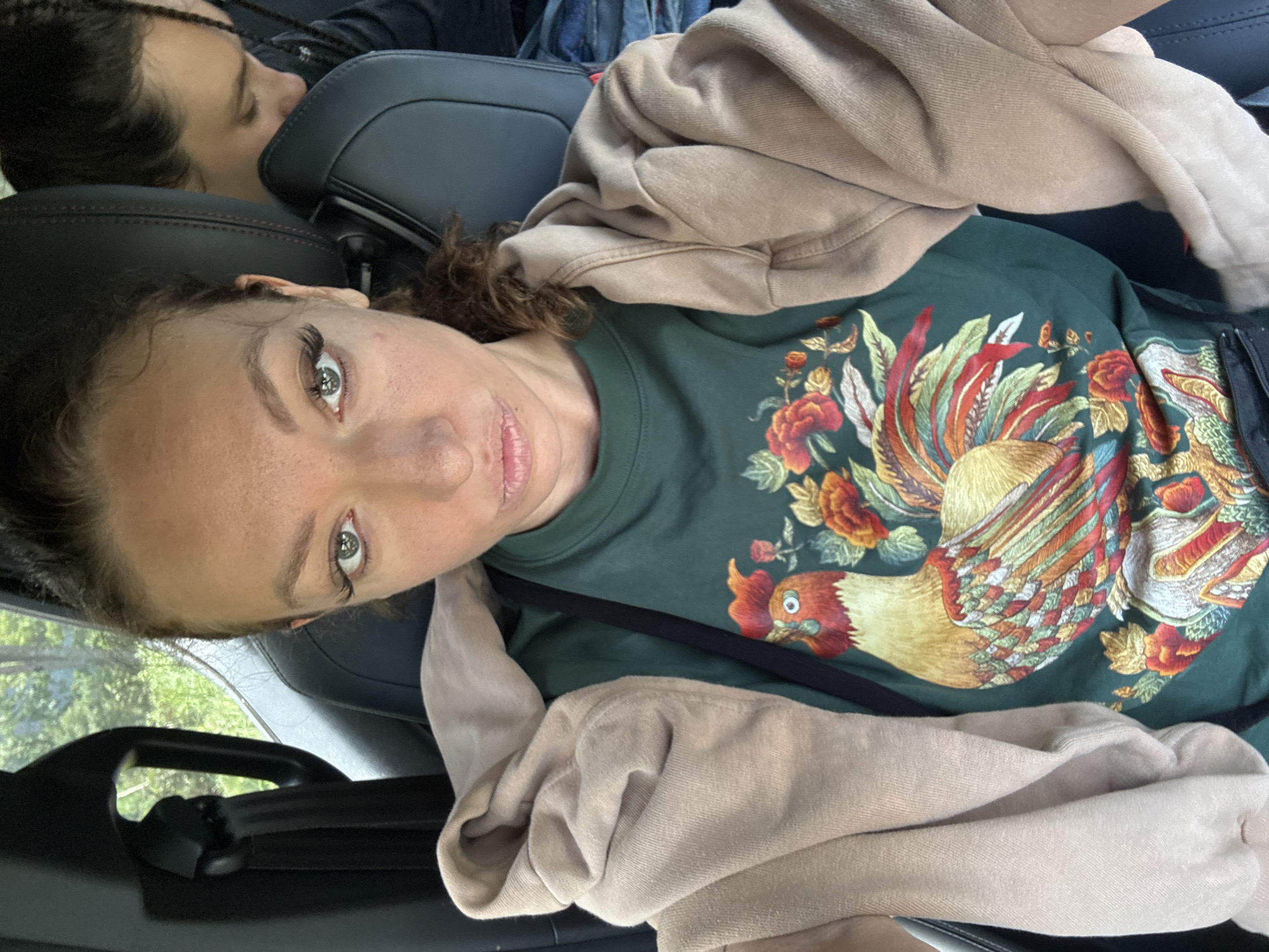 A woman with curly brown hair and green eyes is seated in a car, wearing a green t-shirt with a colorful rooster design and a beige sweatshirt. A second person is visible in the background, partially obscured by the car seat. The car appears to be in transit. The location is not discernible as Hoàng Su Phì Commune, Vietnam.