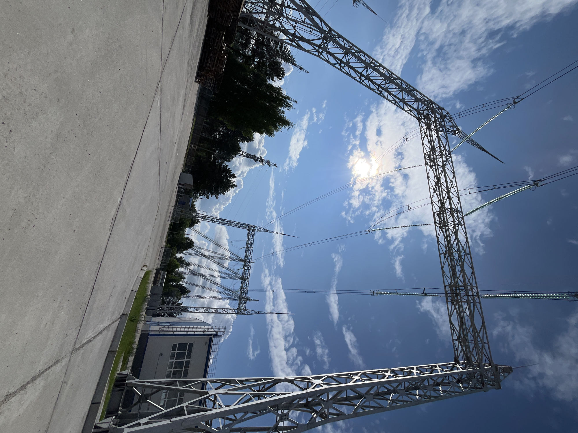 A wide-angle, low-angle outdoor daytime shot captures a complex electrical substation under a partly cloudy blue sky. The foreground is a paved concrete area with visible expansion joints. To the left, a stack of wooden pallets is piled high. Dominating the frame are large, intricate metal structures supporting high-voltage power lines. These towers are composed of crisscrossing metal beams, creating a lattice-like appearance. The power lines themselves are thick and extend across the sky, supported by insulators. In the mid-ground, more power line towers and trees are visible. On the right side of the image, a light-colored industrial building with multiple windows and a metal ladder on its side is present. The sun is bright and partially obscured by clouds, creating a lens flare effect. The overall scene suggests a functional industrial site, likely part of the energy infrastructure of Chaikovsky, Russia.