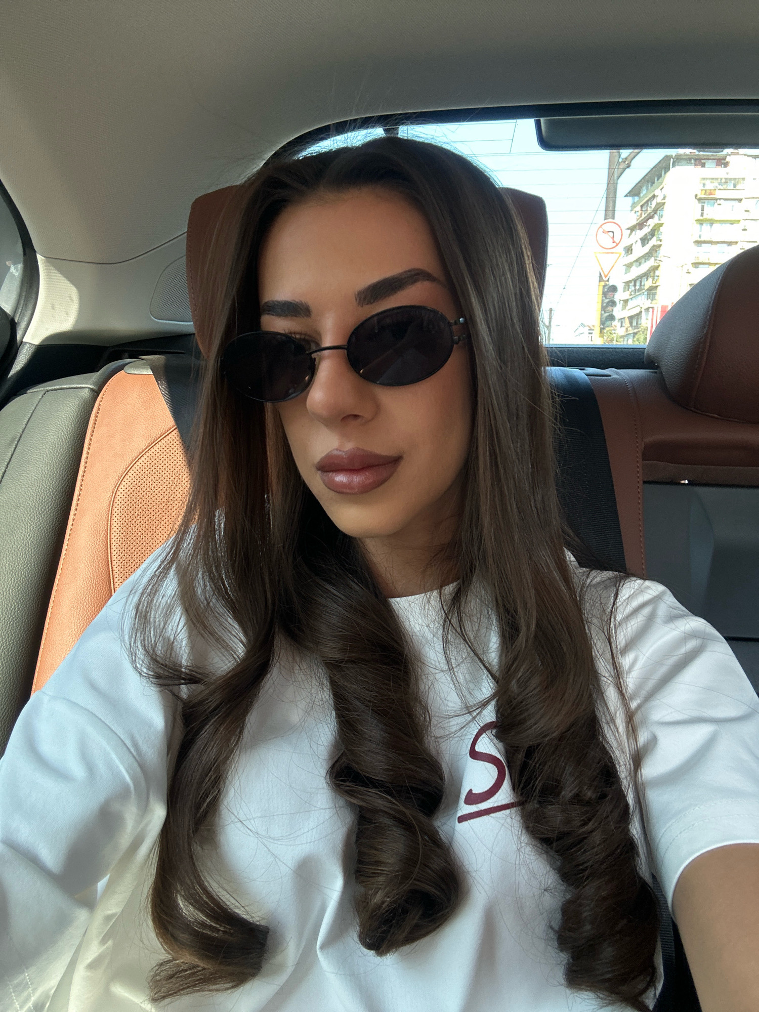 A young woman with long, dark, wavy hair and dark sunglasses is sitting in the back seat of a car. She is wearing a white t-shirt with a maroon letter "S" and a line underneath on the left chest. The car's interior appears to be leather, with brown and black seats. Outside the car window, a street scene in the city of Sofia, Bulgaria, is visible, featuring buildings and a traffic light. The image is a selfie, taken from a slightly high angle.