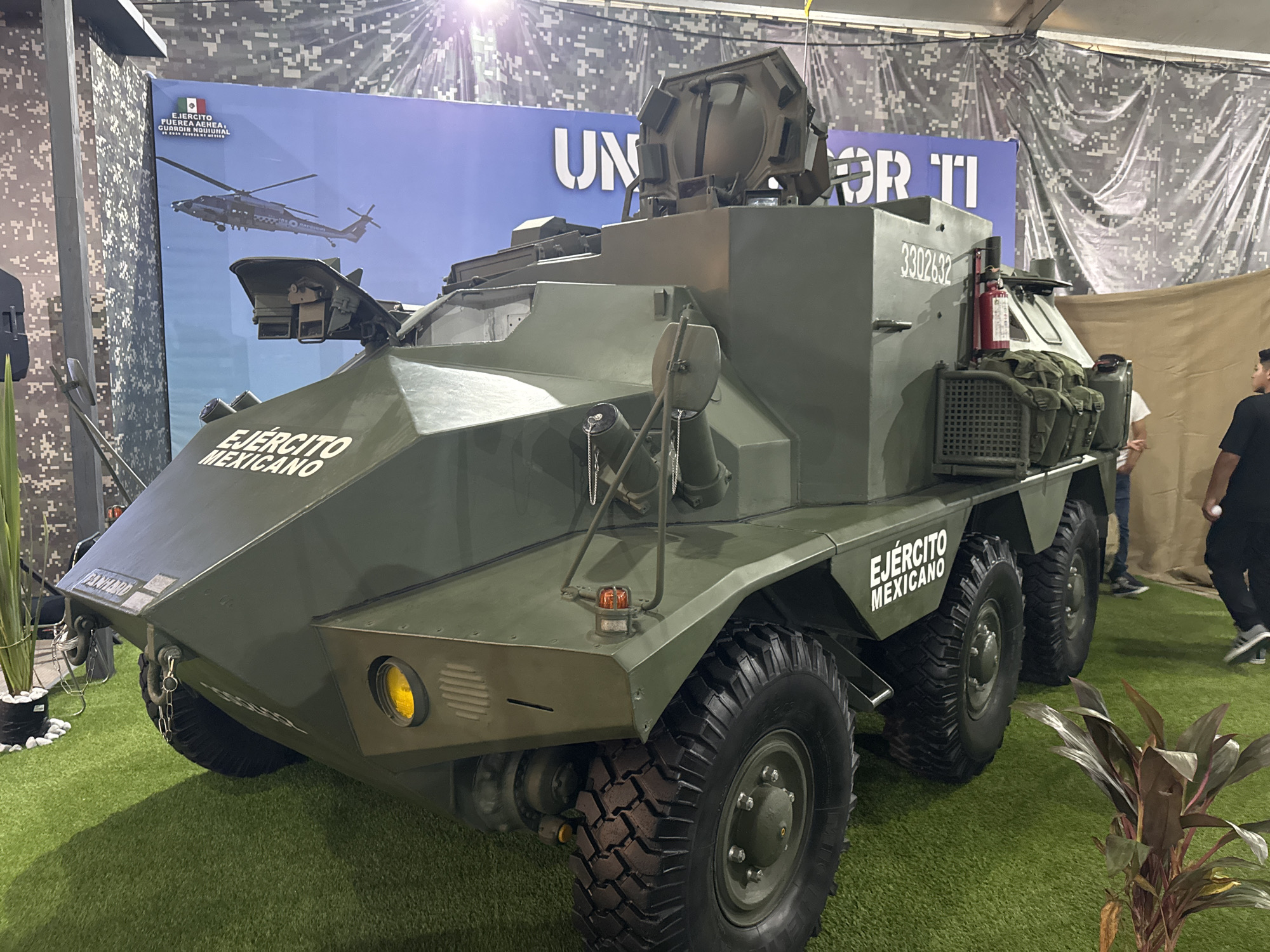 The image shows a Mexican Army armored vehicle on display. The vehicle is painted olive green and has "EJÉRCITO MEXICANO" (Mexican Army) written on its sides. It is a 6x6 wheeled vehicle. The background includes a banner with "UN" and "TI" visible, as well as an image of a helicopter. The vehicle is situated on artificial green grass. The setting appears to be an exhibition or indoor event, possibly in Puebla, Mexico, given the "EJÉRCITO MEXICANO" markings. People are visible in the background. The number 3302632 is visible on the vehicle. A fire extinguisher is also mounted on the vehicle. There are potted plants to the side of the vehicle.