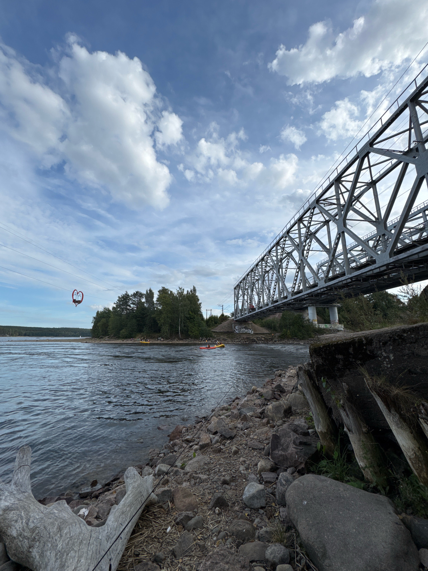A wide-angle, low-angle shot captures a river and a large, industrial-style bridge in Losevo, Russia. The sky is partly cloudy with white cumulus clouds scattered against a blue backdrop. The river dominates the lower half of the frame, with its surface rippling under the overcast sky. The rocky shore in the foreground is littered with driftwood and loose stones.
To the left, a heart-shaped cable car hangs suspended over the water by thick cables, its bright red frame a stark contrast to the muted tones of the landscape. Further in the distance, a wooded shoreline is visible with a line of evergreen trees. Several kayaks are dotted on the river, with people paddling.
The massive steel bridge, a prominent feature of the scene, stretches across the upper right of the image, its intricate latticework structure casting shadows on the water. The bridge appears to be a railway bridge, with tracks visible along its surface. Concrete pylons support the bridge in the distance. On the embankment leading up to the bridge, graffiti is visible. The overall scene depicts a blend of natural and industrial elements, with human activity evident in the kayaks on the river and the cable car.