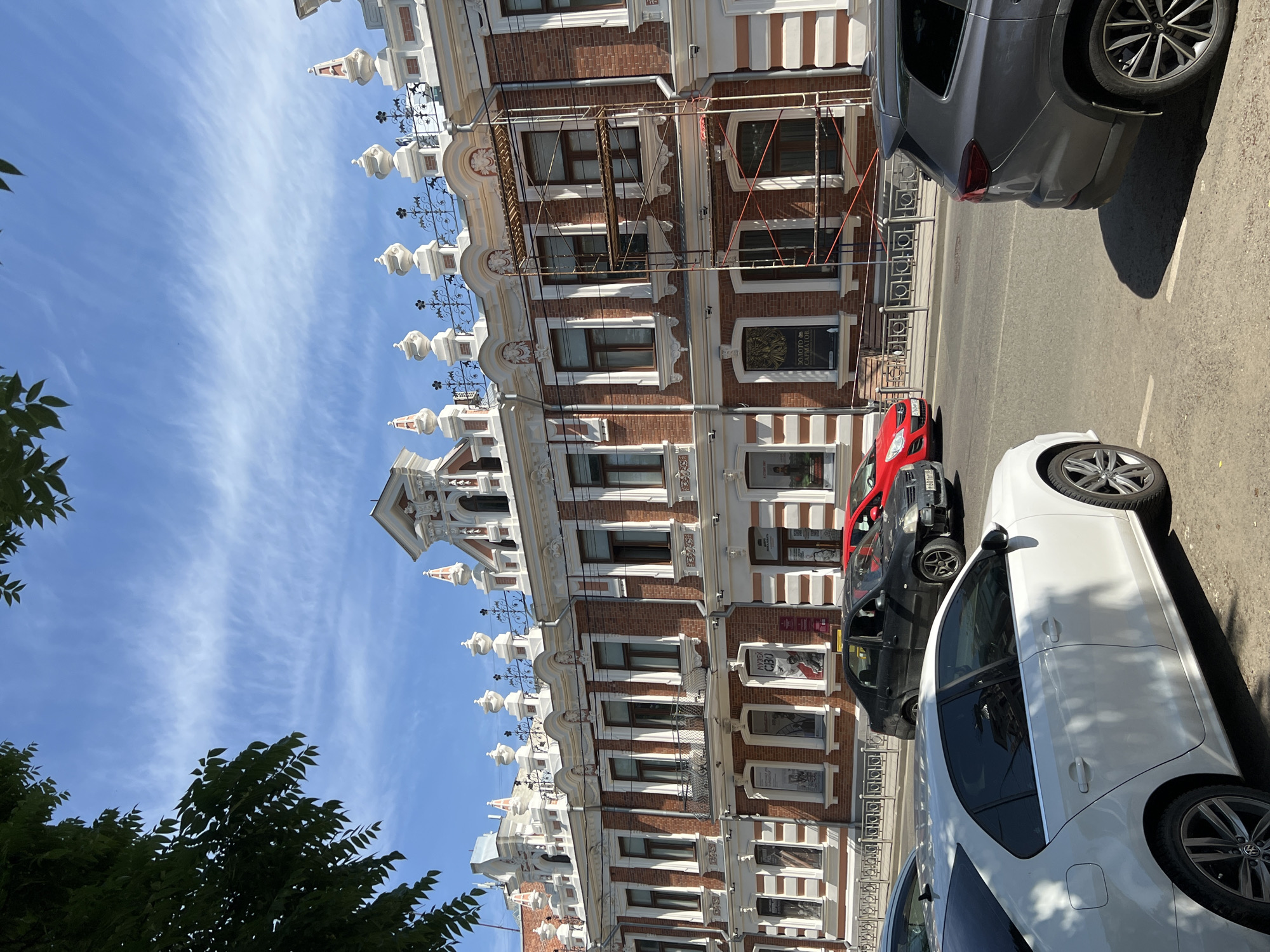 An exterior, eye-level shot captures a historic brick building with white ornate trim and decorative roofline accents under a blue sky with wispy clouds. Scaffolding is visible on the right side of the building, suggesting ongoing renovation. The building has multiple windows and some posters displayed in them. In the foreground, several cars are parked on a street, including a white sedan on the left, a dark sedan and a red car in the middle, and a dark gray SUV on the right. The scene depicts a street in Krasnodar, Russia.