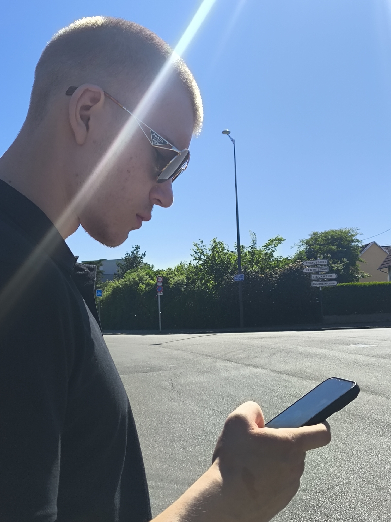 The image shows a fair-skinned young man in Hoenheim, France, holding a smartphone. He wears Prada sunglasses and a dark shirt. He stands on a grey asphalt road, and the background includes vegetation, a lamppost, street signs indicating directions to Strasbourg, La Wantzenau, Bischheim, and Hoenheim Centre, and a glimpse of a building. The image is taken in bright sunlight, creating lens flare.
