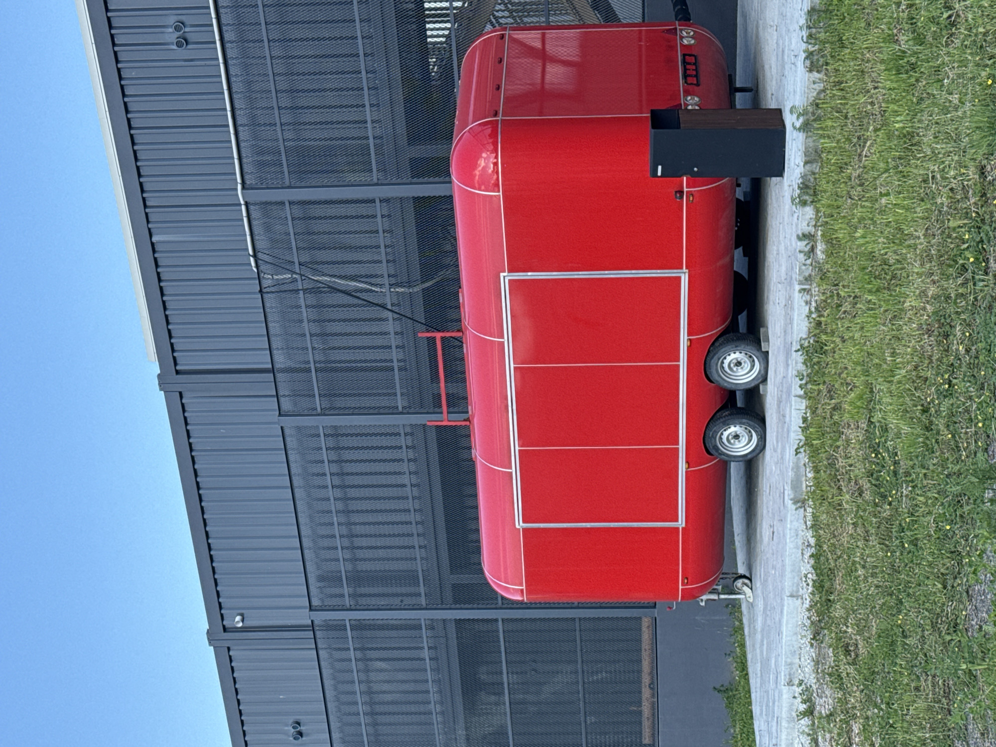 A bright red trailer, possibly a food truck or mobile stand, is parked outdoors on a paved surface next to a grassy area. The trailer has a curved front and a rectangular door with a metallic frame. It is supported by two wheels visible on the right side. A dark, box-like attachment is affixed to the side of the trailer near the front. A metal ladder is partially visible attached to the side of the trailer. In the background, a modern building with a dark, corrugated metal facade and a latticed metal screen runs along the side. The sky above is clear and blue. The overall impression is of an outdoor commercial setup, possibly in an industrial or commercial area. The lighting suggests it is daytime, likely sunny. There are no people visible in the image. There is some text on the back of the trailer, reading "000" above what appears to be three dials or buttons.