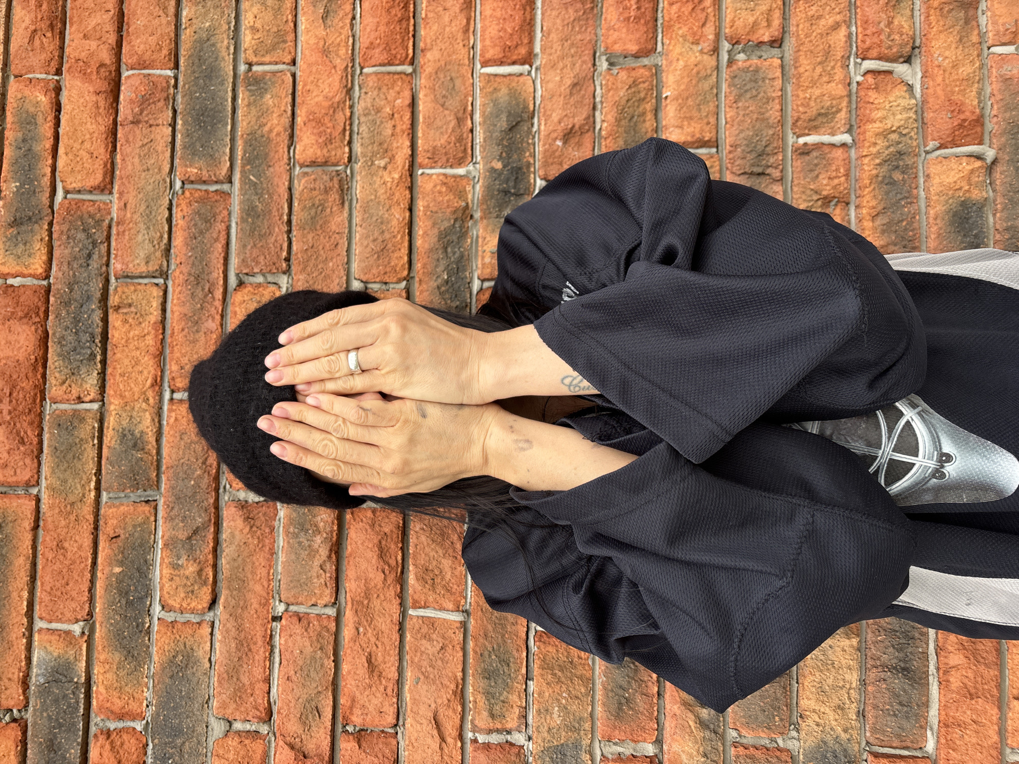 An overhead photograph captures a person lying on a brick surface, oriented vertically within the frame. Their head is positioned towards the left edge, and their body extends towards the right. Their face is entirely obscured by both hands, which cover their eyes and forehead. The individual wears a dark, likely black or navy blue, long-sleeved garment made of a textured, possibly athletic fabric. Dark hair is visible around their hands.
A silver-colored ring is worn on the ring finger of the left hand. A tattoo, featuring partially visible cursive letters and a symbolic design, is present on the person's left forearm. The clothing includes a light-colored stripe, possibly white or light grey, visible on the right side of the frame. A metallic object with a circular design and intersecting lines is partially visible near the lower right, appearing to be underneath or part of the garment.
The person is lying on a brick surface that forms the entire background. The bricks are predominantly reddish-orange with variations in shade, including some darker, almost burnt-looking bricks. The mortar lines are light grey and have a somewhat rustic appearance. The brick pattern runs vertically throughout the image frame.
The activity suggests the person is intentionally hiding their face, which could convey emotions such as shyness, exhaustion, distress, or a playful gesture. Bright, even lighting indicates daytime conditions. The brick surface serves as a location cue, suggesting an outdoor setting like a patio or walkway, or an indoor space with brick flooring, relevant to the context of Chaikovsky, Russia. The text on the tattoo is not fully legible.