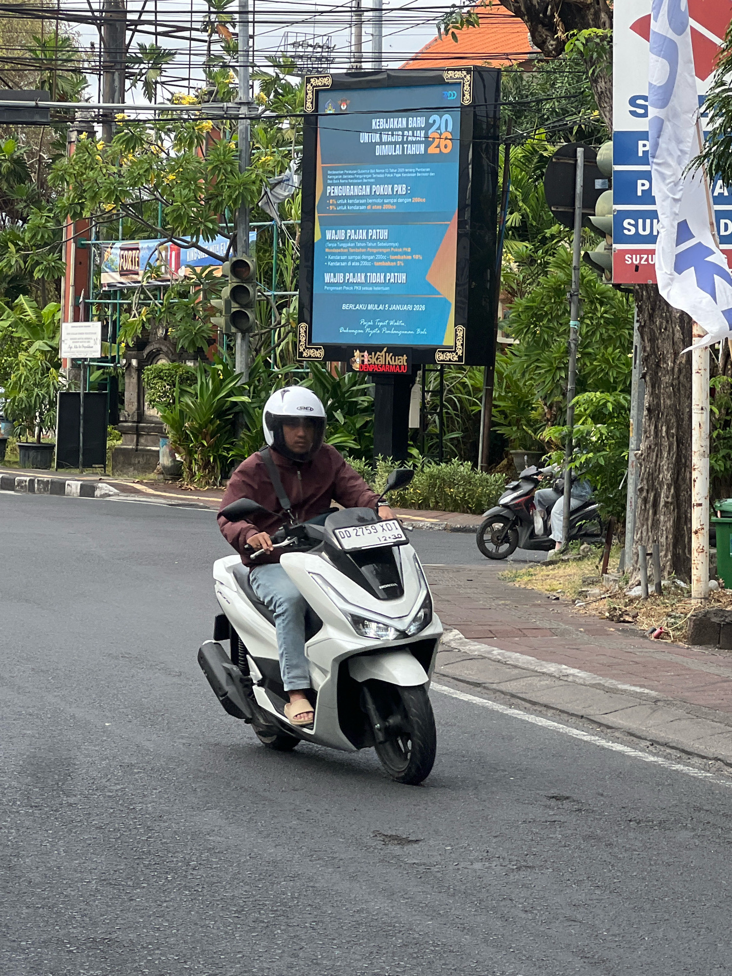 The image depicts a street scene in Denpasar, Indonesia, with a person riding a white scooter. The rider is wearing a maroon jacket, blue jeans, and a white helmet. The scooter has a license plate that reads "DD 2759 XDI". In the background, there is a large billboard with Indonesian text, traffic lights, lush green trees, and other vehicles. The road is paved, and there is a sidewalk with some debris. Another scooter is parked on the side of the road. A white flag with blue lettering is also visible on the right side of the image.