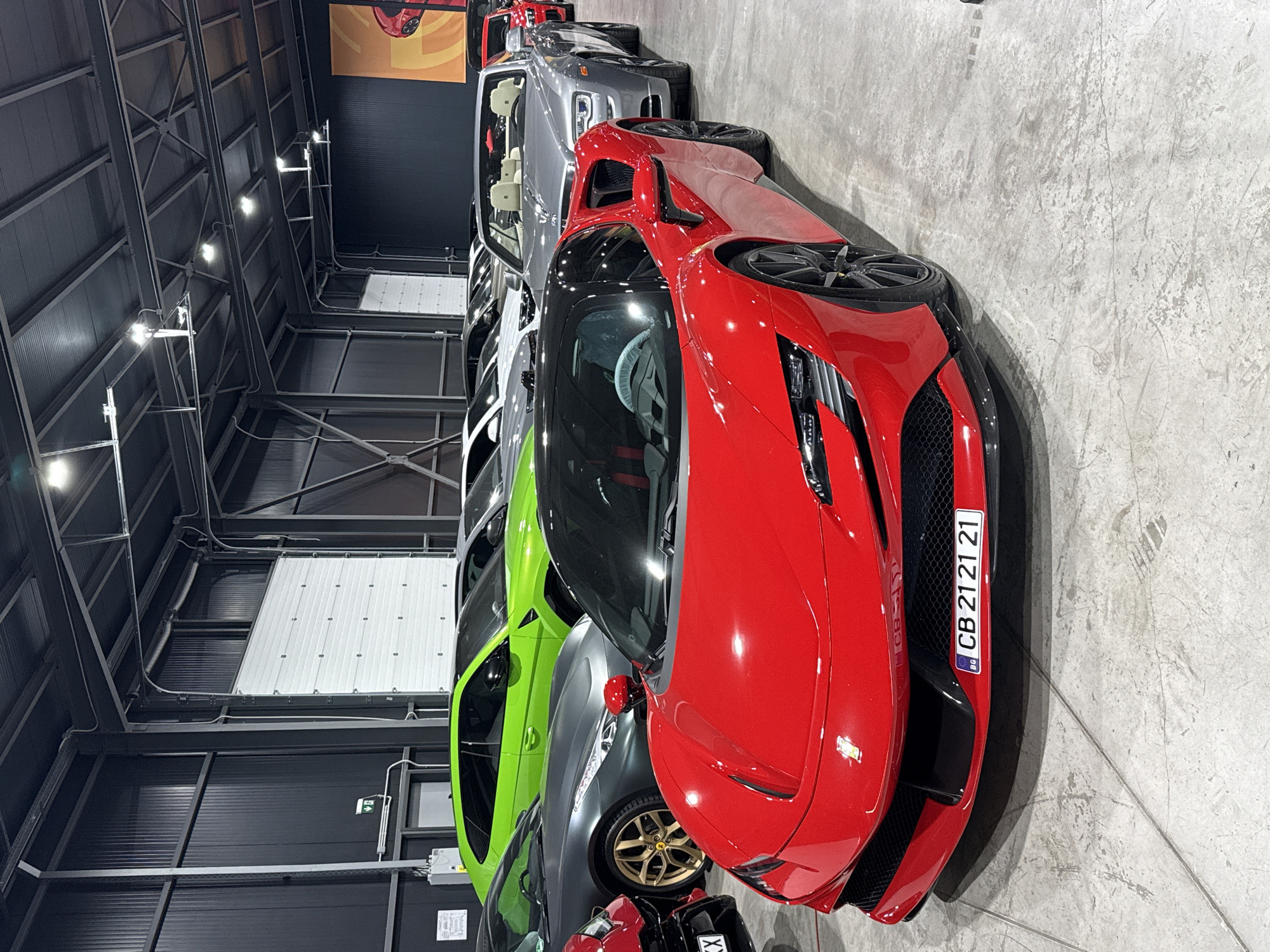A red Ferrari SF90 Stradale is parked indoors, likely in a dealership or private collection, with its front prominently displayed. The car has a black roof and a license plate reading "BG CB 2121 21". Behind it, a line of other luxury vehicles is visible, including a bright green Lamborghini SUV and a silver convertible, possibly a Rolls-Royce. The environment appears to be a modern, well-lit building with a concrete floor and industrial-style walls and ceiling, featuring large white garage doors. The image does not contain any people. This scene is consistent with the automotive showrooms often found in commercial areas of Plovdiv, Bulgaria.