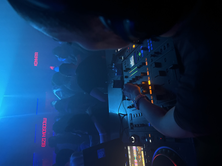 The image depicts an indoor nightclub or event venue in Moscow, Russia, characterized by low light and a pervasive blue-cyan illumination with atmospheric haze. In the foreground, a person, likely a DJ, is partially visible from behind, facing away from the camera. This individual wears headphones and their right arm, adorned with a metallic bracelet/watch, is positioned over a DJ mixer, actively adjusting controls. The mixer features multiple illuminated knobs, faders, and screens, one displaying a waveform and tempo information (106 BPM). To the left of the mixer, a laptop (possibly a MacBook Pro with a "BP" sticker) is open on the DJ console. In the blurred background, a dense crowd of indistinct individuals is present, illuminated by the blue light. Two red neon signs are visible against the dark wall: one reading "BIZI MOSCOW" and the other displaying "КОМНАТА" (KOMNATA), which translates to "ROOM" in Russian.