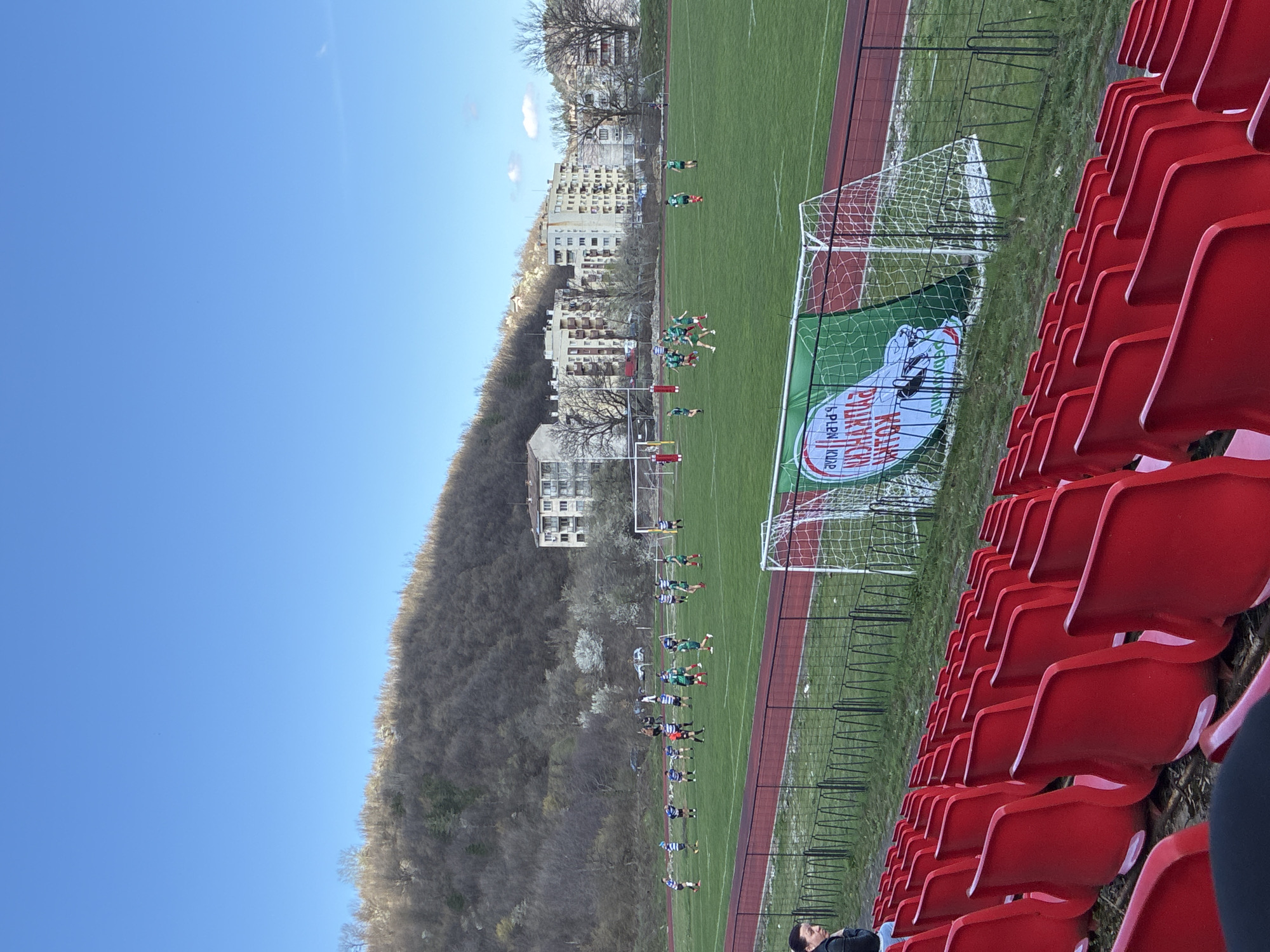 The image shows a soccer game being played on a green field in Berkovitsa, Bulgaria. In the background, there are buildings and hills, possibly the Balkan Mountains. The field is lined with red running tracks. In the foreground, red stadium seats are visible, indicating the presence of a spectator area. Some people are present in the stadium. Players dressed in green and white or blue and white uniforms are on the field, possibly in the middle of a game, with two teams lined up. There is a soccer goal with a promotional banner displayed. The sky is blue.