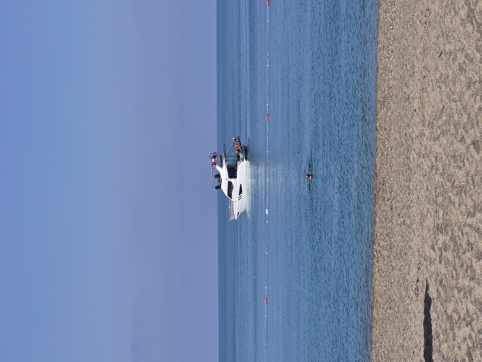 The image shows a beach scene in Alanya, Turkey. A white yacht is anchored in the blue sea, near a string of buoys. Several people are on the yacht, and one person is swimming in the water nearby. The shoreline consists of sand and pebbles, with a shadow visible on the beach. The sky is clear and blue.