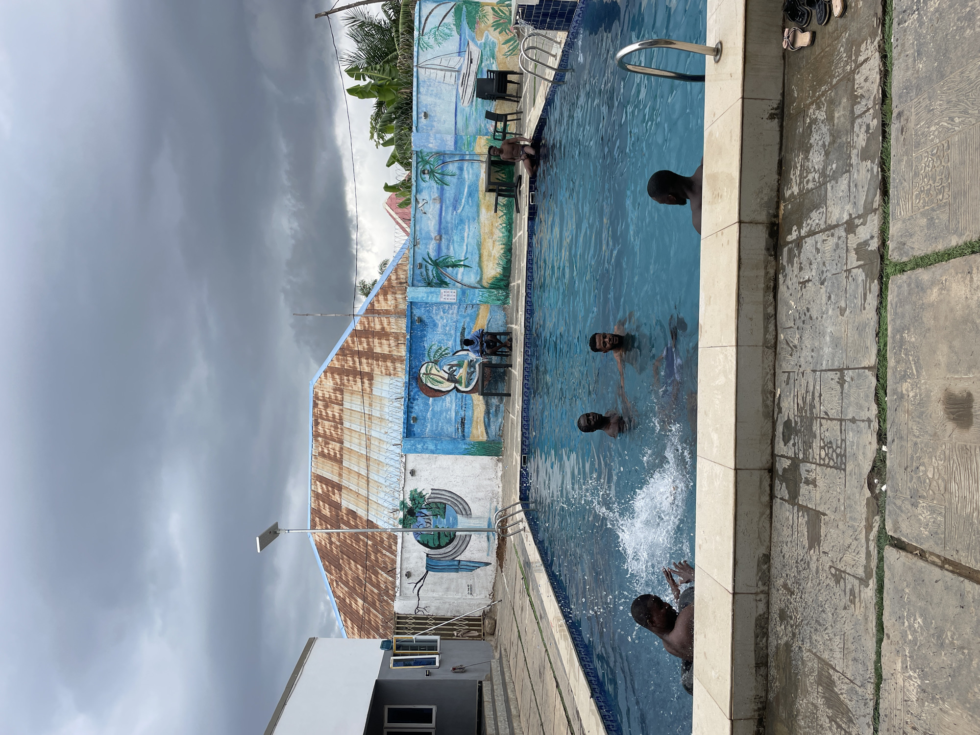 The image depicts a swimming pool scene outdoors, likely in Akwanga, Nigeria, given the visual characteristics. Several people are in the pool, their heads and shoulders visible above the water's surface. One person is seated on the edge of the pool. The pool is rectangular with blue tiles inside. A mural depicting a beach scene with palm trees and a sailboat adorns the wall adjacent to the pool. The mural has a weathered appearance. A building with a corrugated metal roof and a white painted facade is behind the mural, which suggests a residential or recreational area. The sky is overcast with dark, heavy clouds. The pool deck is made of concrete tiles. A pile of footwear is beside the pool on the deck. A light pole is visible near the building. The water is active.