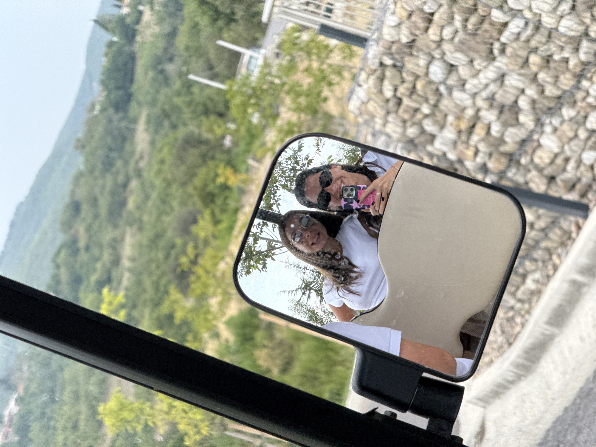 A reflection in a side-view mirror shows two young women taking a selfie in a golf cart. The woman on the right holds a pink-cased smartphone, and both women wear sunglasses and white t-shirts. The mirror reflects a hilly, green landscape with trees and some buildings in the distance. The golf cart is on a paved surface next to a stone wall and a curb. The overall impression is of a sunny day in a rural or semi-rural setting, possibly a resort or vineyard in Sighnaghi, Georgia, known for its scenic beauty.