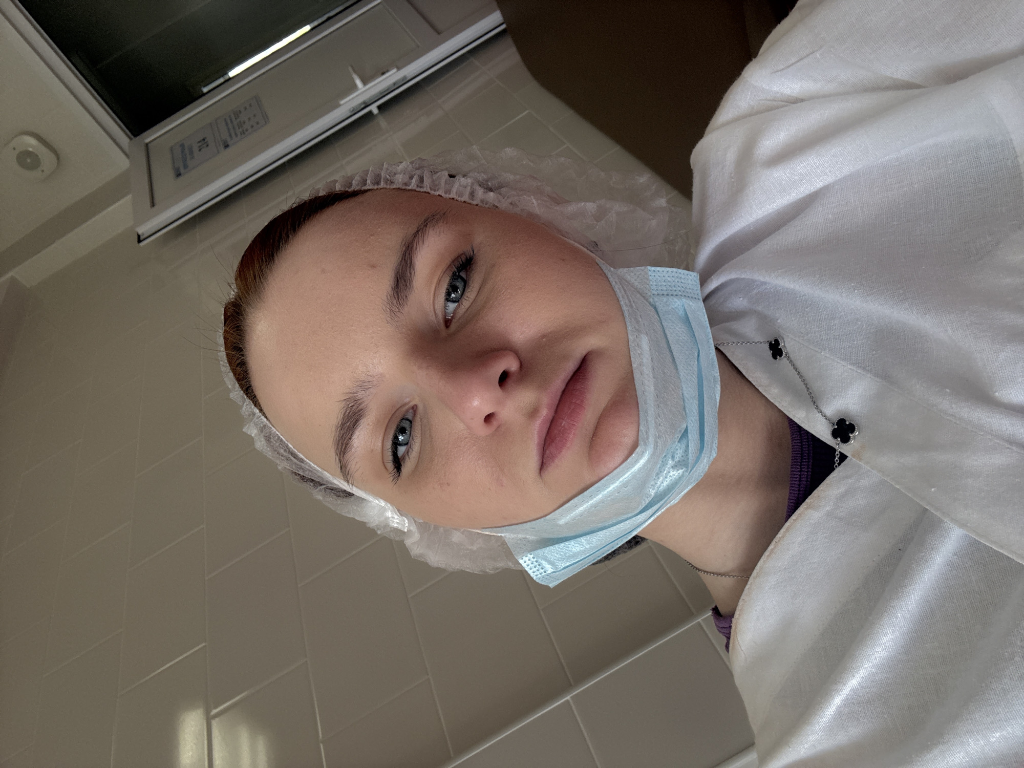 A medium close-up shot shows a woman wearing a white hairnet and a light blue surgical mask pulled down below her mouth. She is also wearing a white lab coat over a dark shirt, and a necklace with a clover-shaped pendant is visible. The woman has light skin, blue eyes, and reddish-brown hair. The background consists of light-colored tiled walls and a fluorescent light fixture. A partial view of a piece of furniture is seen at the right-upper corner of the image. Based on the style and environment, it is likely an indoor medical or healthcare setting. There is no indication that the image was taken in Saint Petersburg, Russia, and no location information is provided.