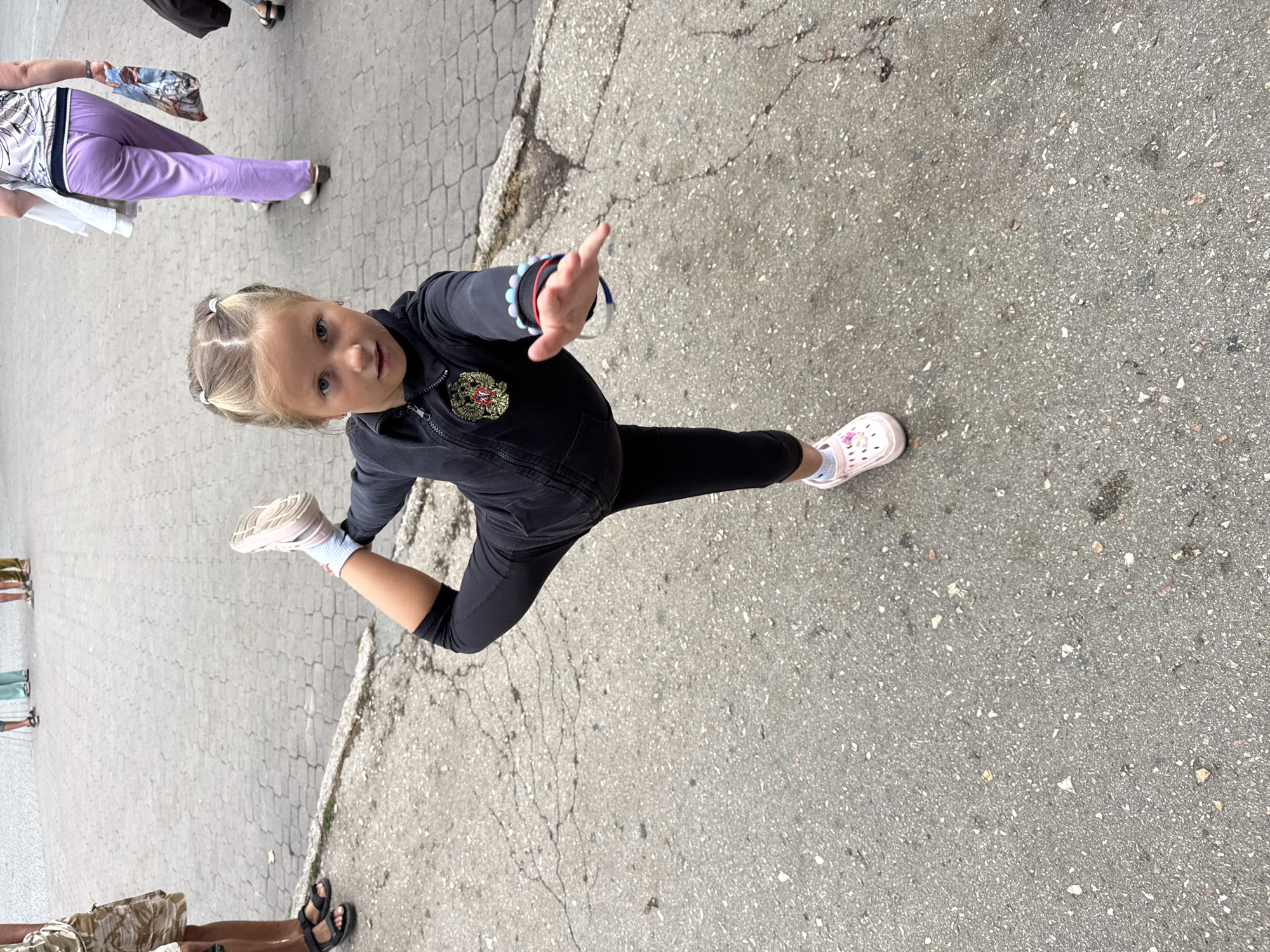 A young girl with blonde hair styled in pigtails is performing a dance pose on a paved area. She is wearing a black zip-up jacket with a crest on the left chest, black leggings, white socks, and pink clogs with a floral design. Her right leg is bent and extended behind her, and her left arm is outstretched forward, palm open. The ground is a mix of asphalt and paver stones. In the background, several people are walking, including a woman in light purple pants and a patterned top, and a person wearing patterned shorts. The scene appears to be outdoors in a public space. This image was taken in Inkerman, Ukraine.