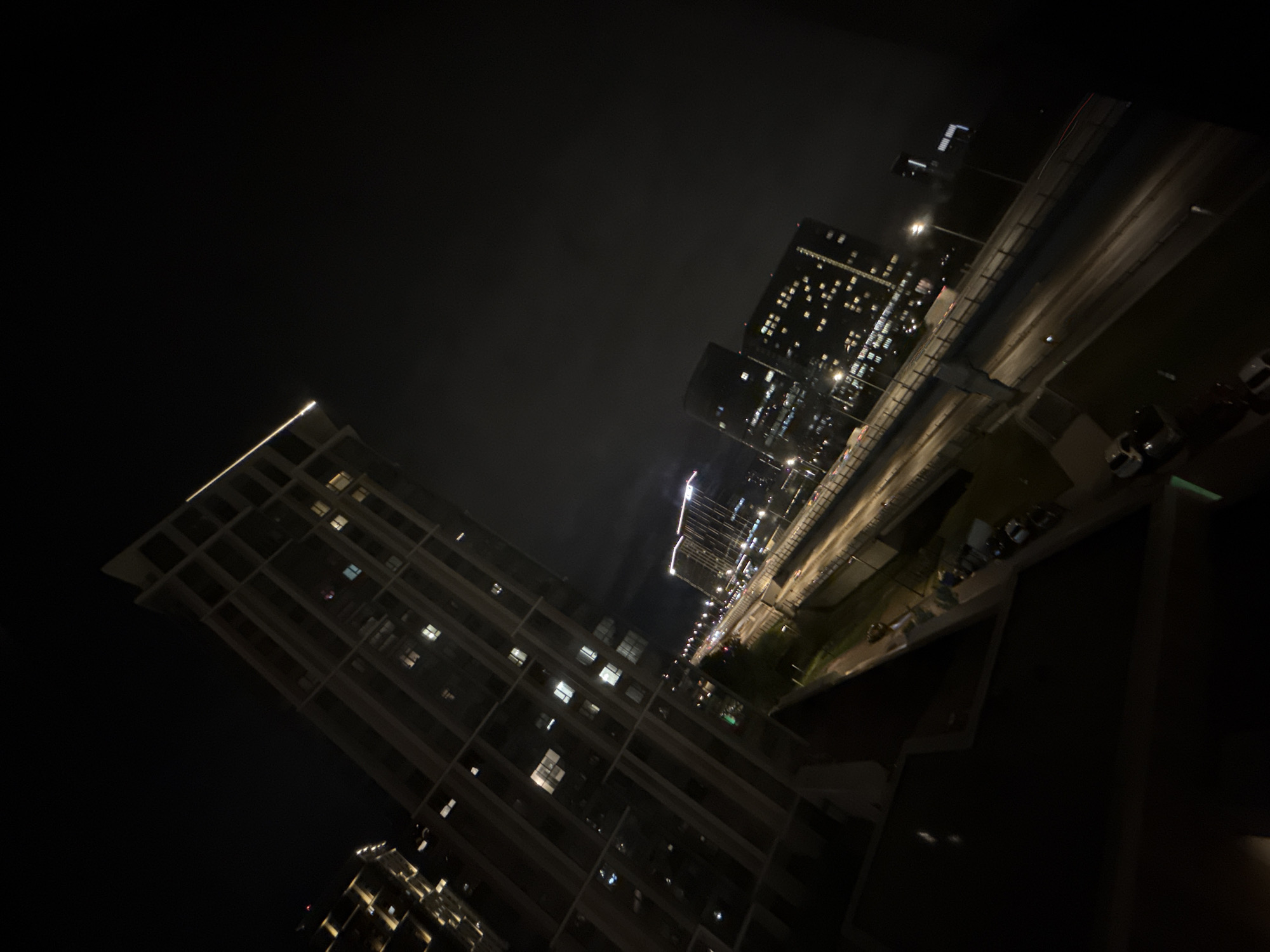The image depicts a night view in Khimki, Russia, featuring multiple buildings and a section of road. A tall building with a visible grid-like exterior and lighted windows is prominent in the foreground. To the right, another building with densely lit windows is seen alongside a road with streetlights and cars. The sky is dark, suggesting a nighttime setting. The photo appears to be taken from an elevated position, likely a high floor of a building.