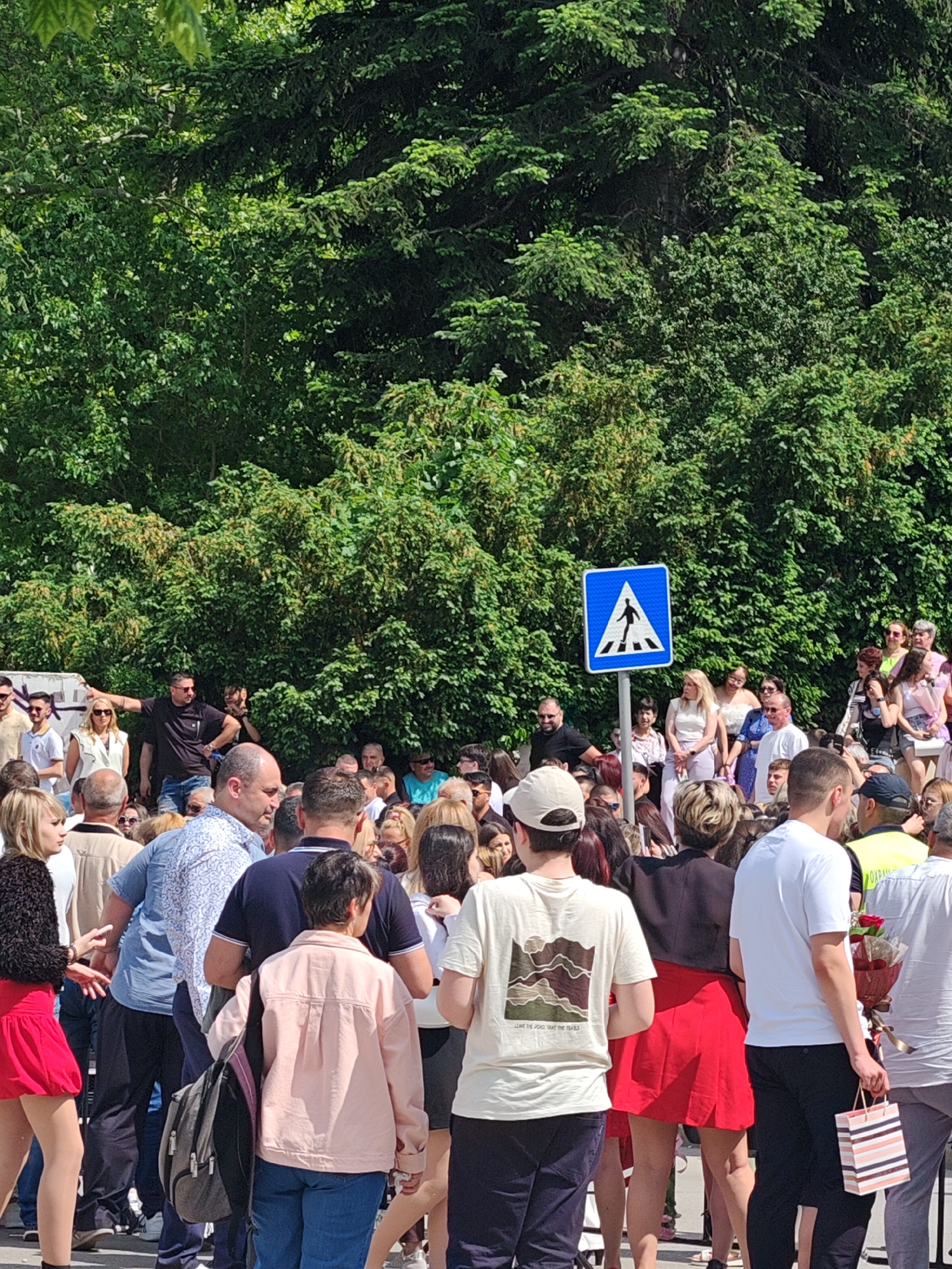 A large group of people are gathered in what appears to be an urban setting in Shumen, Bulgaria. The crowd includes men and women of various ages, standing near a crosswalk sign. The location is outdoors, with trees forming a backdrop. Some individuals are casually dressed, while others appear to be more formally attired. One person is holding a bouquet of red roses and a striped shopping bag. A few people are wearing sunglasses. Some individuals are facing the camera, while others are looking in different directions, suggesting diverse focuses of attention.